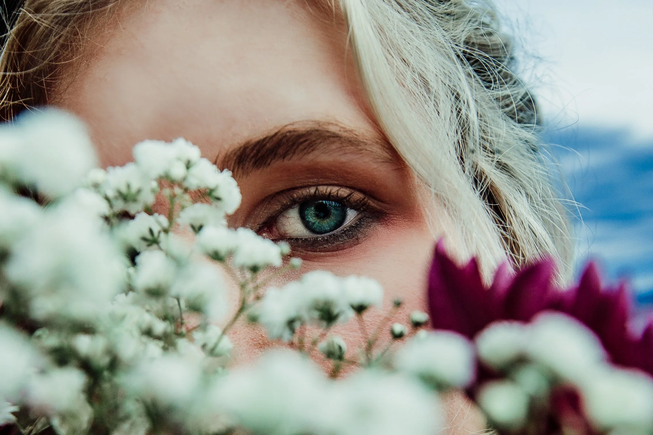 Close-up of a woman's face with striking blue eye, surrounded by white and purple flowers.