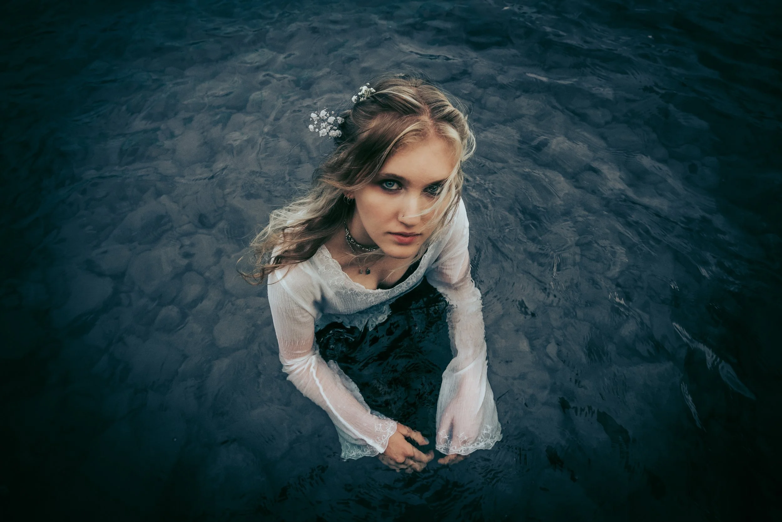 A young woman with blonde hair, blue eyes, and wearing jewelry, is standing in dark water, looking up at the camera with a serious expression.