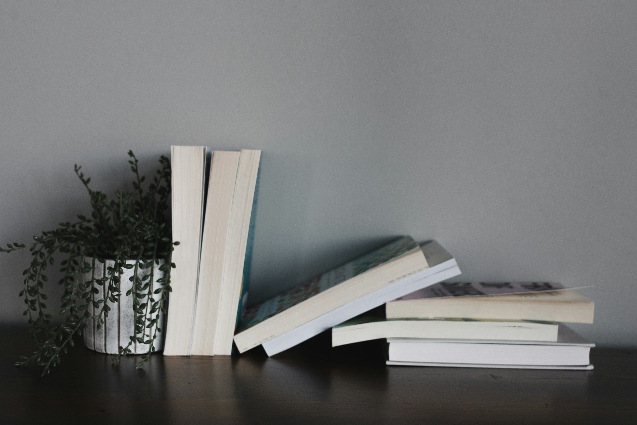 A small potted plant with trailing greenery, a stack of seven books, some leaning, on a dark wooden surface against a light gray wall.