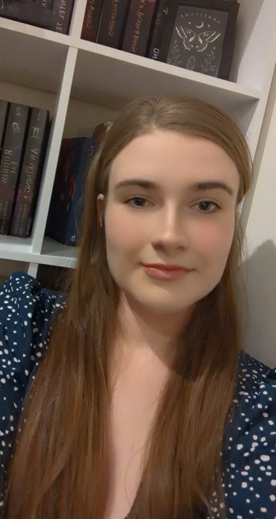 A young woman with long reddish-brown hair, wearing a dark blue shirt with white polka dots, taking a selfie in front of a white bookshelf filled with books.