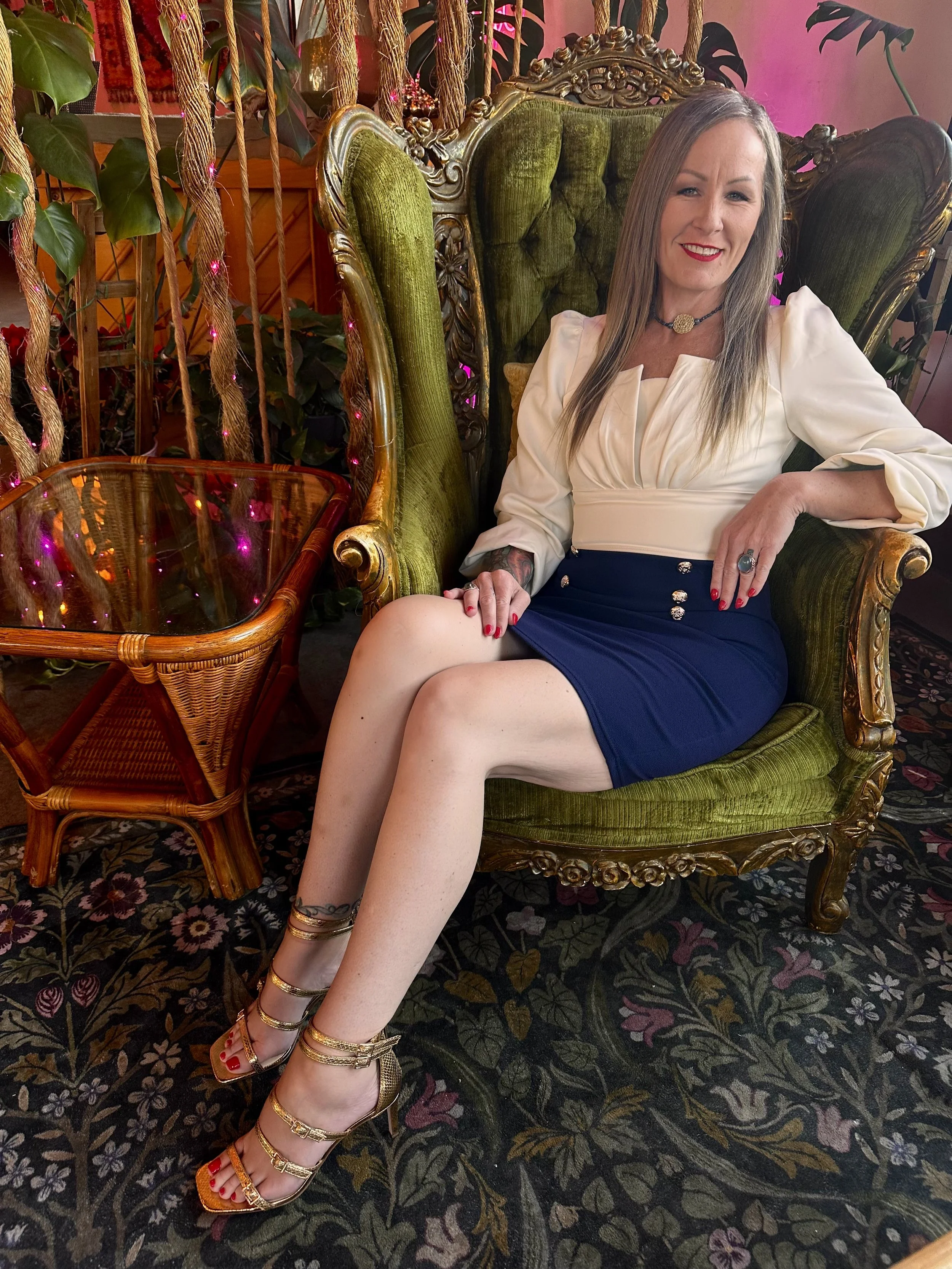 A woman with long hair sitting on a vintage green velvet armchair with wooden carvings, smiling, in a decorated indoor space with plants and pink string lights.