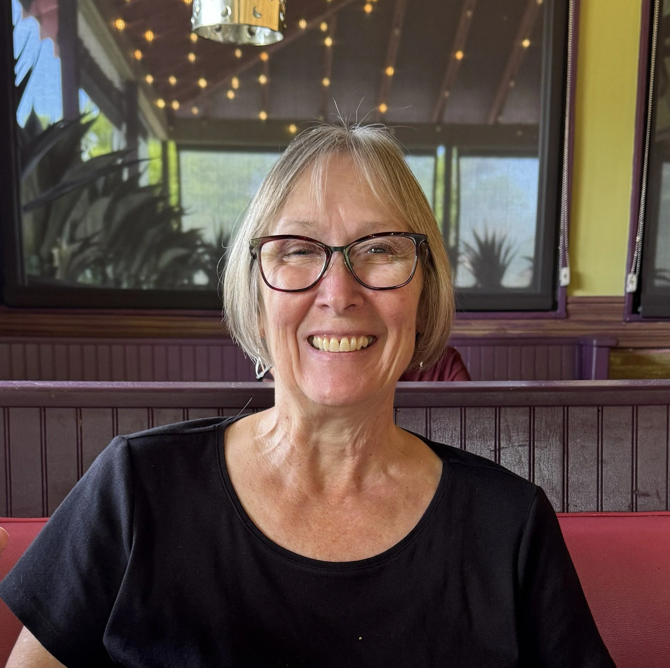A smiling woman with short gray hair, wearing glasses and a black shirt, sitting in a restaurant with wooden decor and a window behind her.