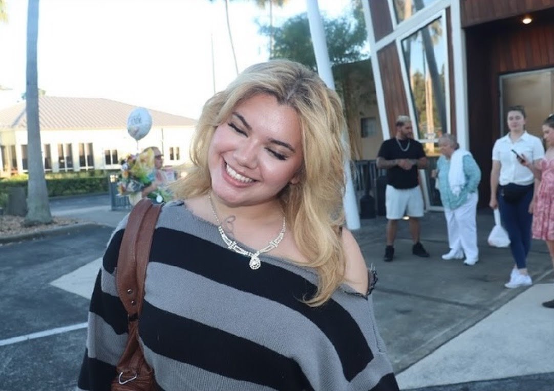 A smiling woman with blonde hair, wearing a black and gray striped top, standing outside a building with other people in the background.