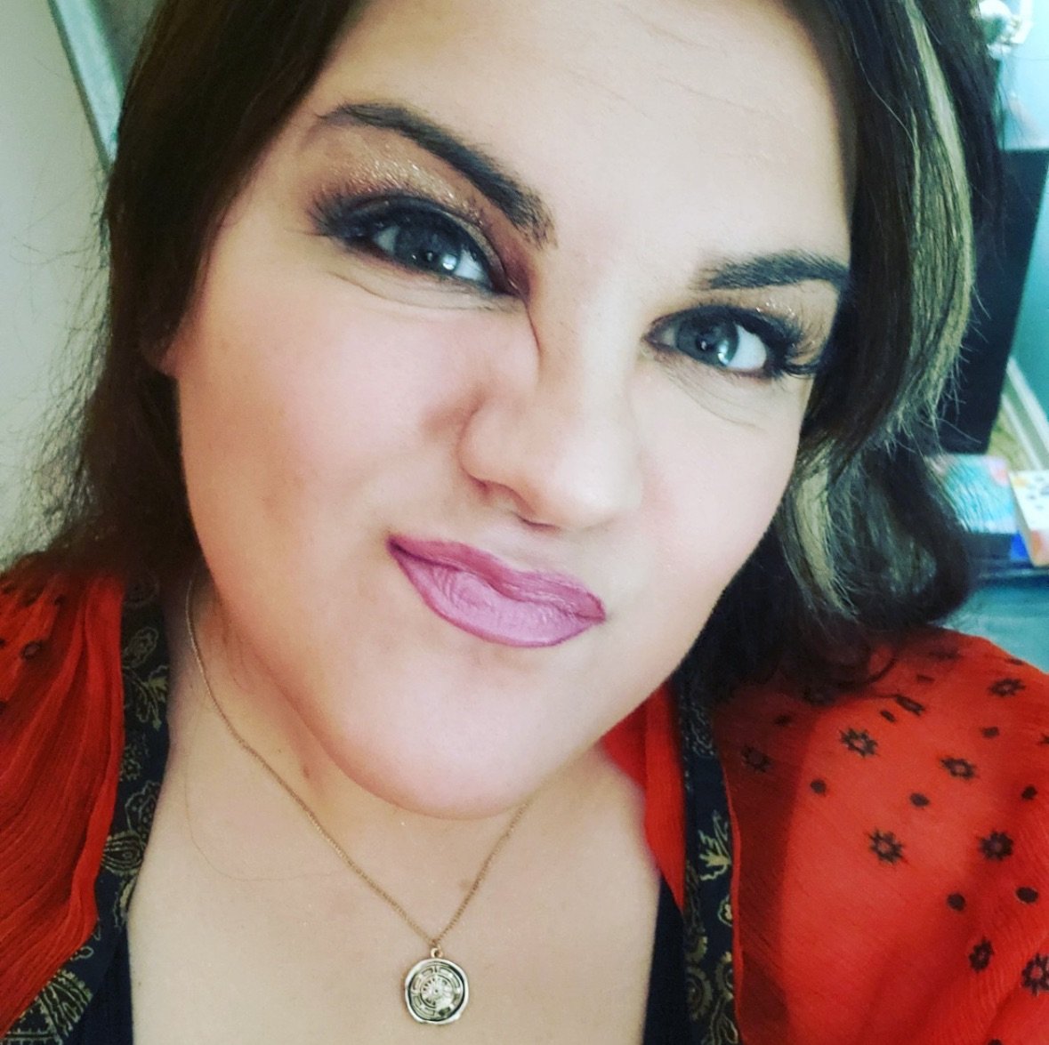 Close-up of a woman with makeup, wearing a red top and a gold pendant necklace, winking and smiling.