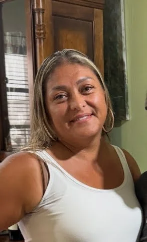 A woman smiling in a room with a green wall and a window with blinds in the background.