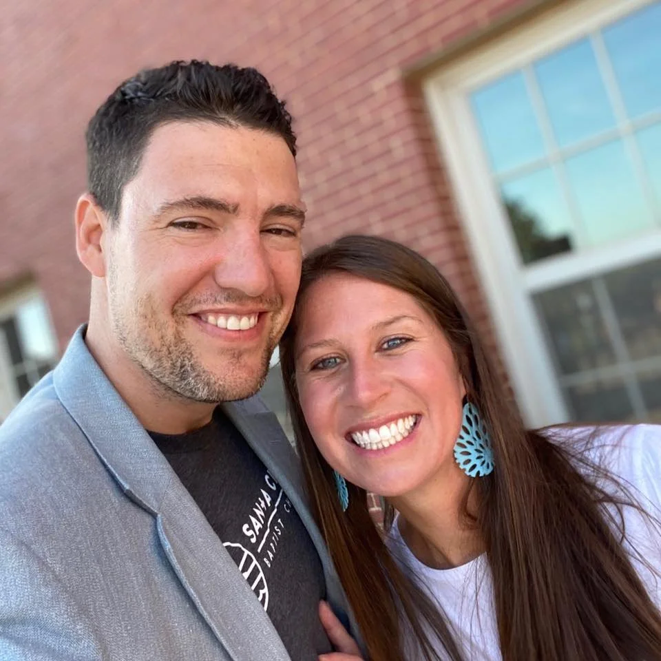 A smiling man and woman taking a selfie together outdoors with a brick building and a window in the background.