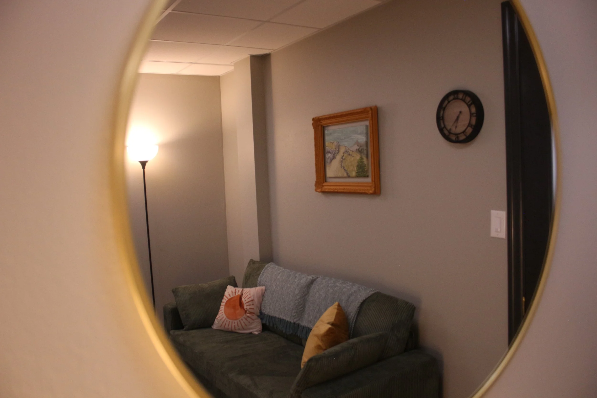 Living room seen through a round mirror, featuring a gray sofa with pillows, a lamp, a framed landscape painting, and a wall clock.