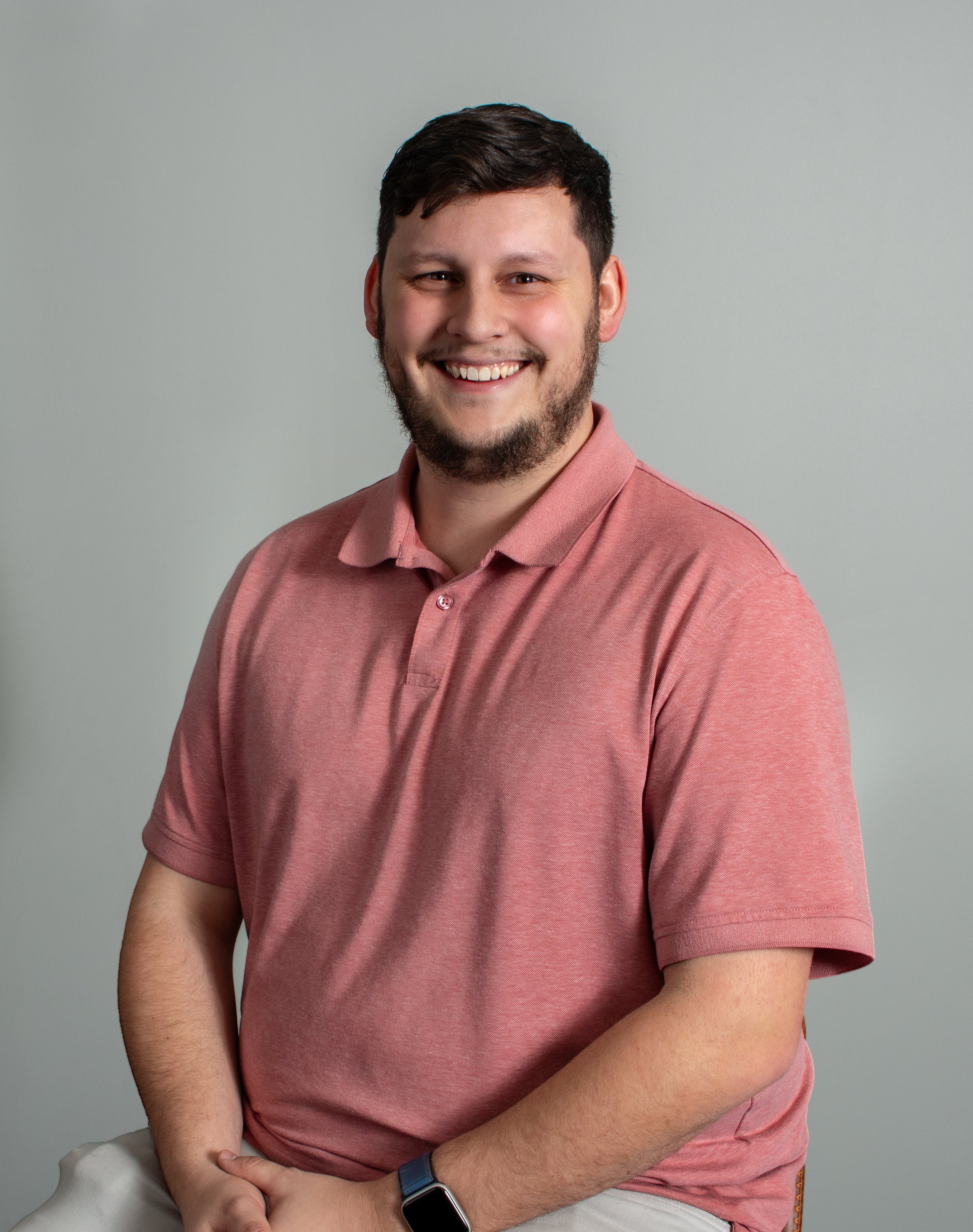 A family of three standing on a lawn in front of a brick house. The woman is holding a young boy, and all three are smiling. The woman wears a navy top and patterned pants, the man wears a color-blocked sweater and beige pants, and the boy wears a checkered shirt and jeans. There are trees and shrubs around them.