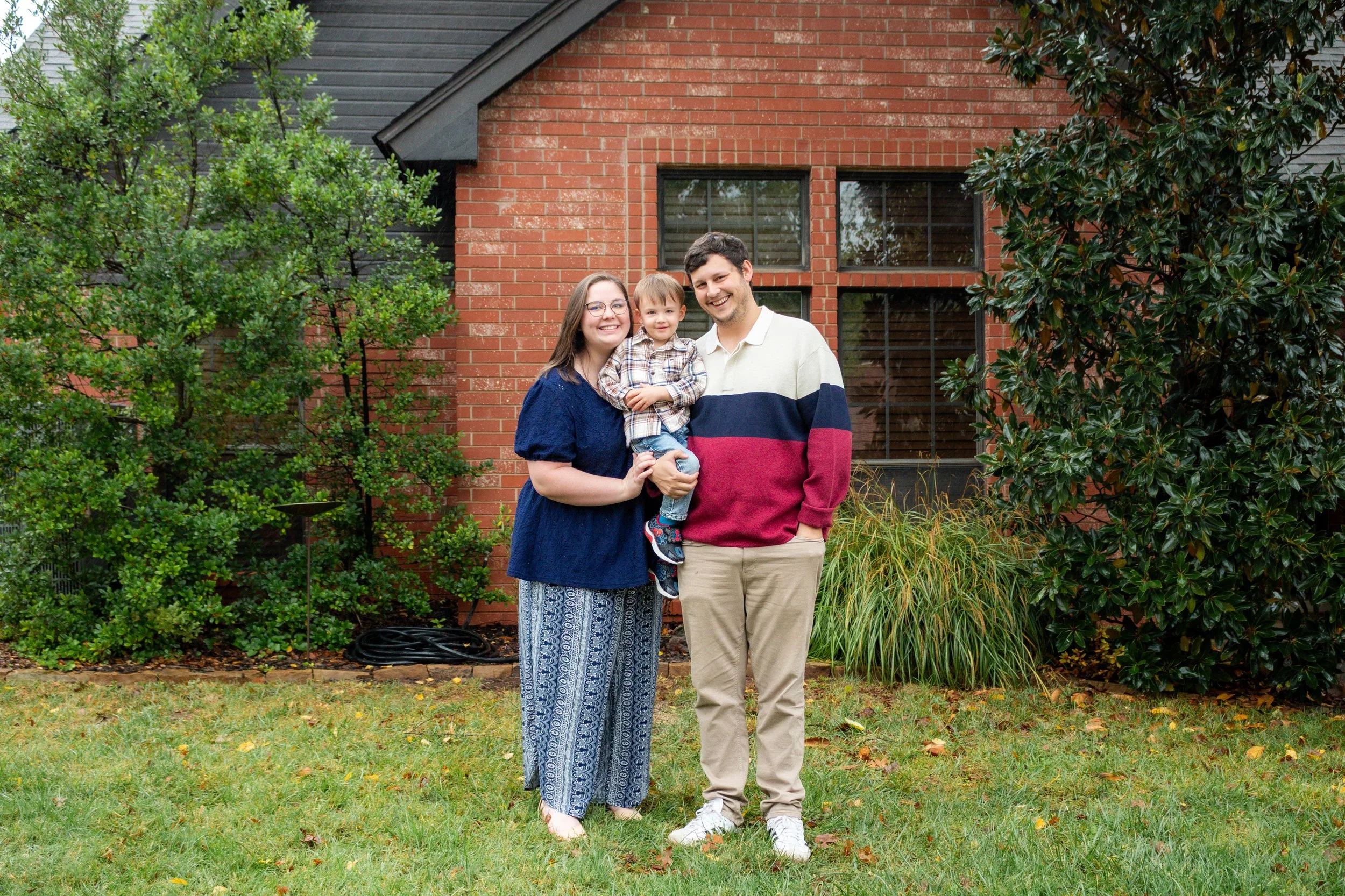A family of three standing on a lawn in front of a brick house. The woman is holding a young boy, and all three are smiling. The woman wears a navy top and patterned pants, the man wears a color-blocked sweater and beige pants, and the boy wears a checkered shirt and jeans. There are trees and shrubs around them.