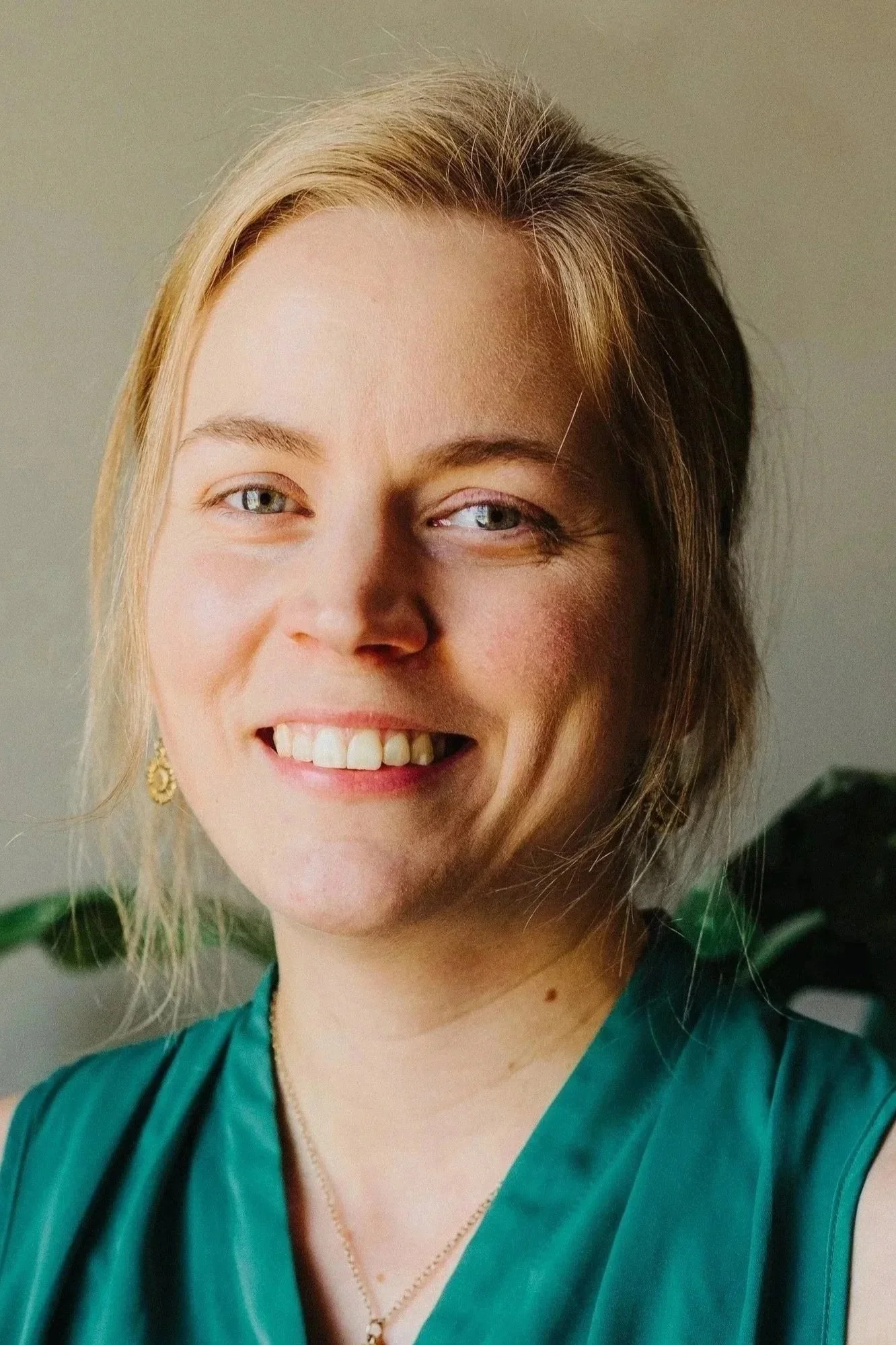 Close-up of a woman smiling, with blonde hair, light blue eyes, wearing a teal dress and a gold necklace, standing next to a green leafy plant.