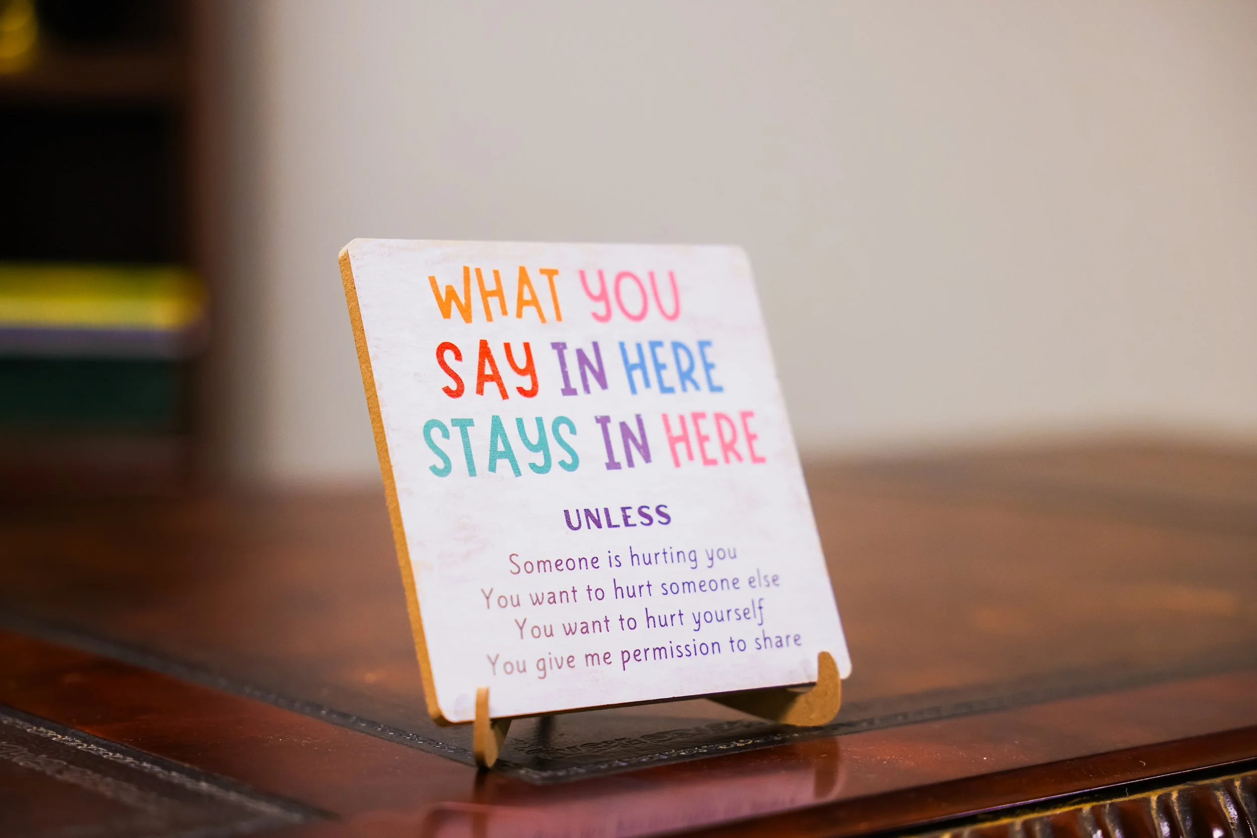 A colorful sign on a small easel with the words 'What you say in here stays in here' written in multicolored letters, placed on a wooden surface with a blurred background.