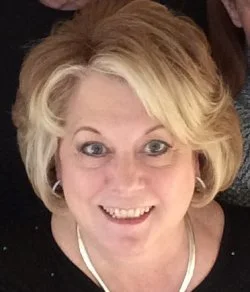 A middle-aged woman with blonde, styled hair, smiling, wearing a black top and a necklace.