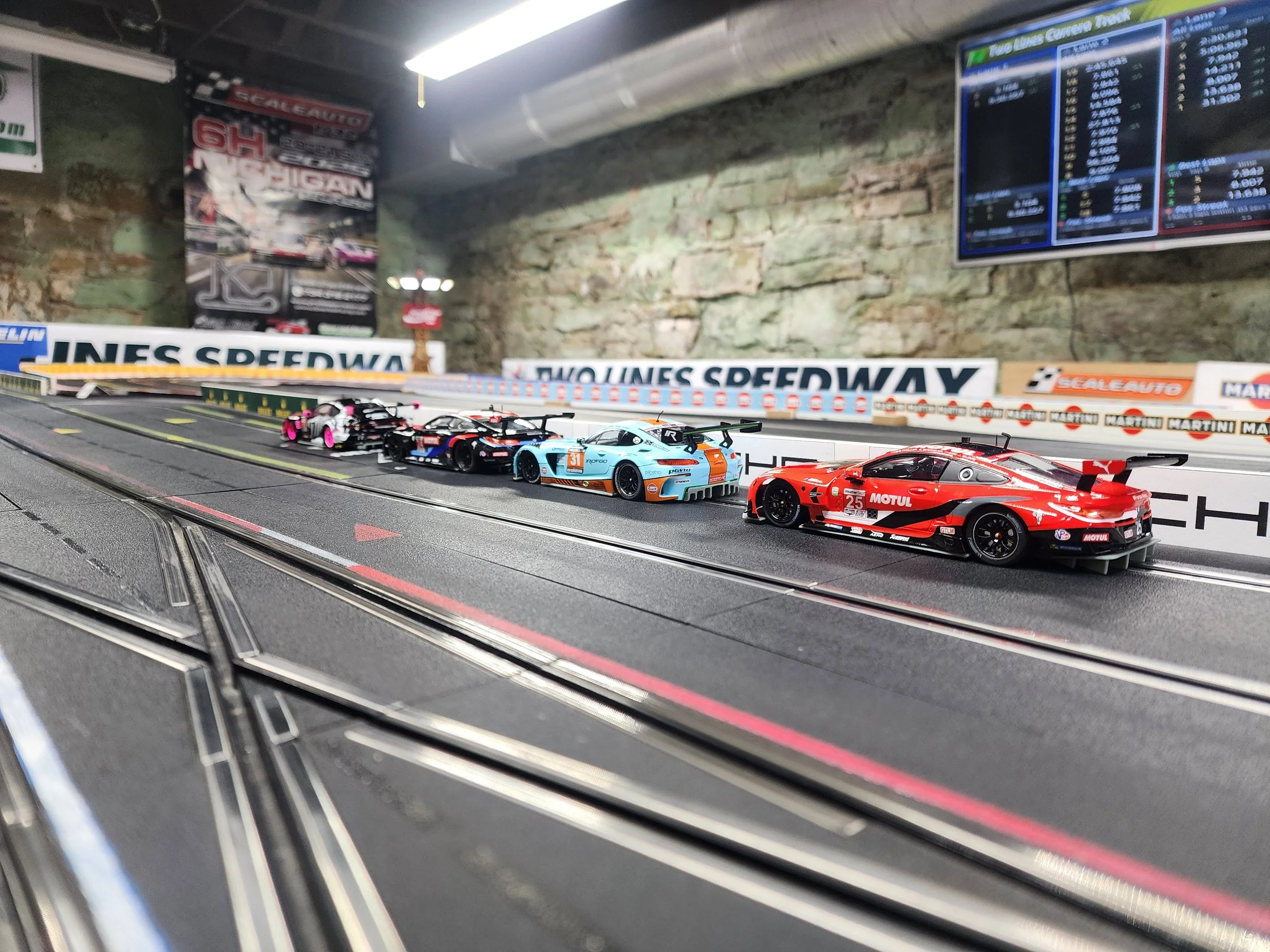 Model race cars on a scale slot car racing track inside a room with a stone wall. The backdrop includes banners and a large screen displaying race information.