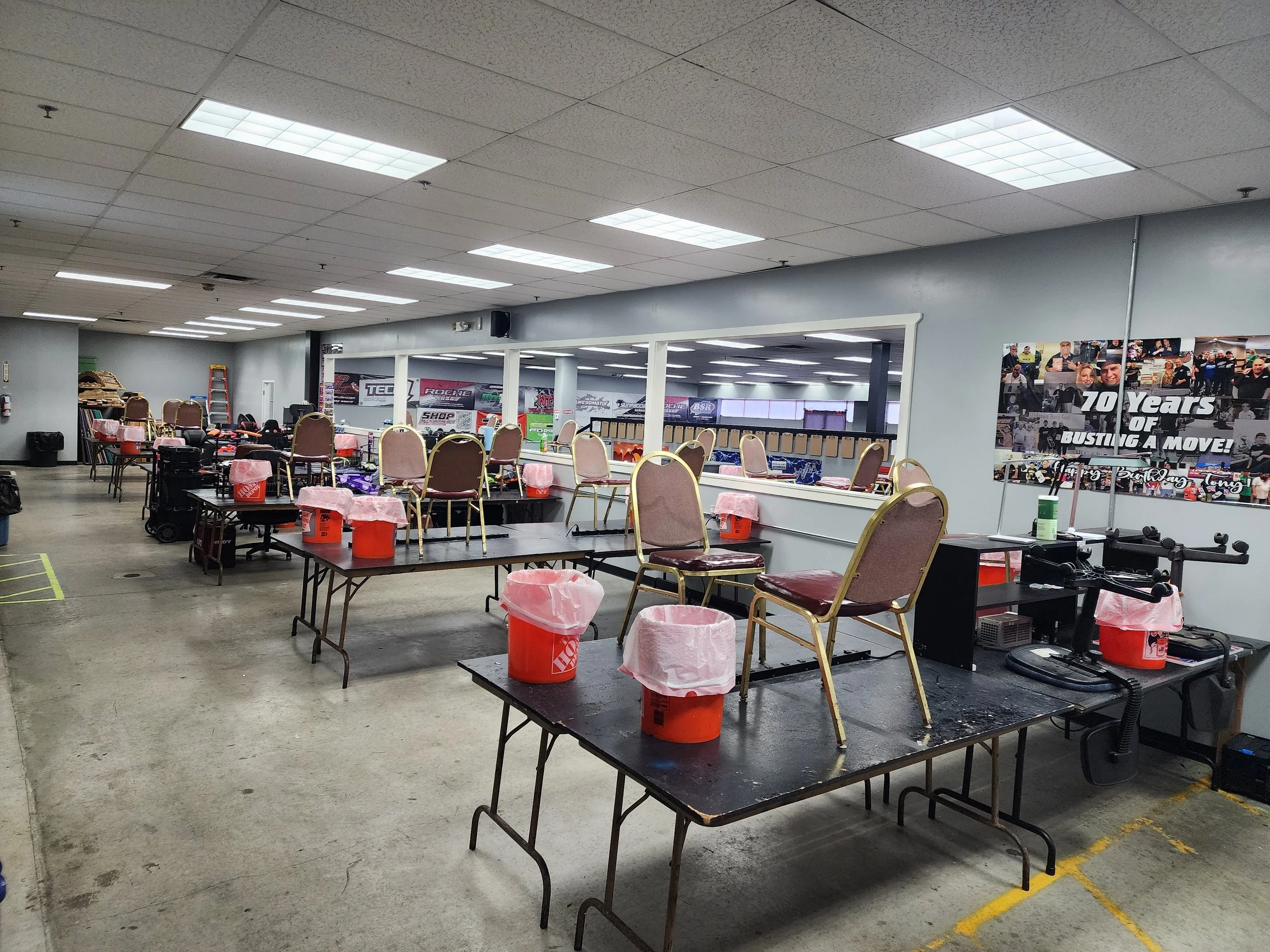 Empty chairs and tables with orange buckets covered in plastic, set up in a large indoor space for a charity event or auction, with photos and banners on the wall.