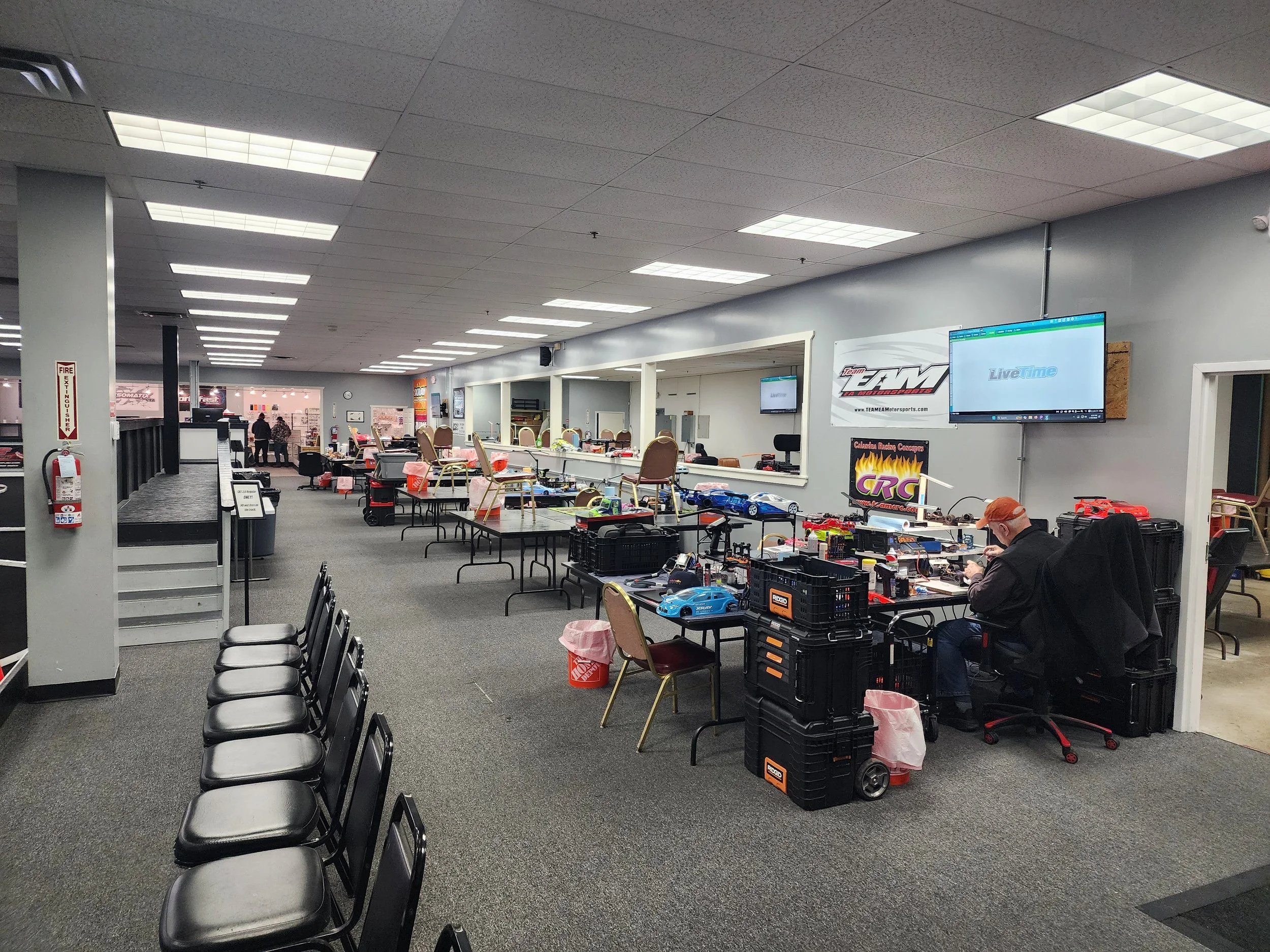 Large indoor space used for remote-controlled car racing, with work tables, chairs, RC cars, and monitors on the wall.