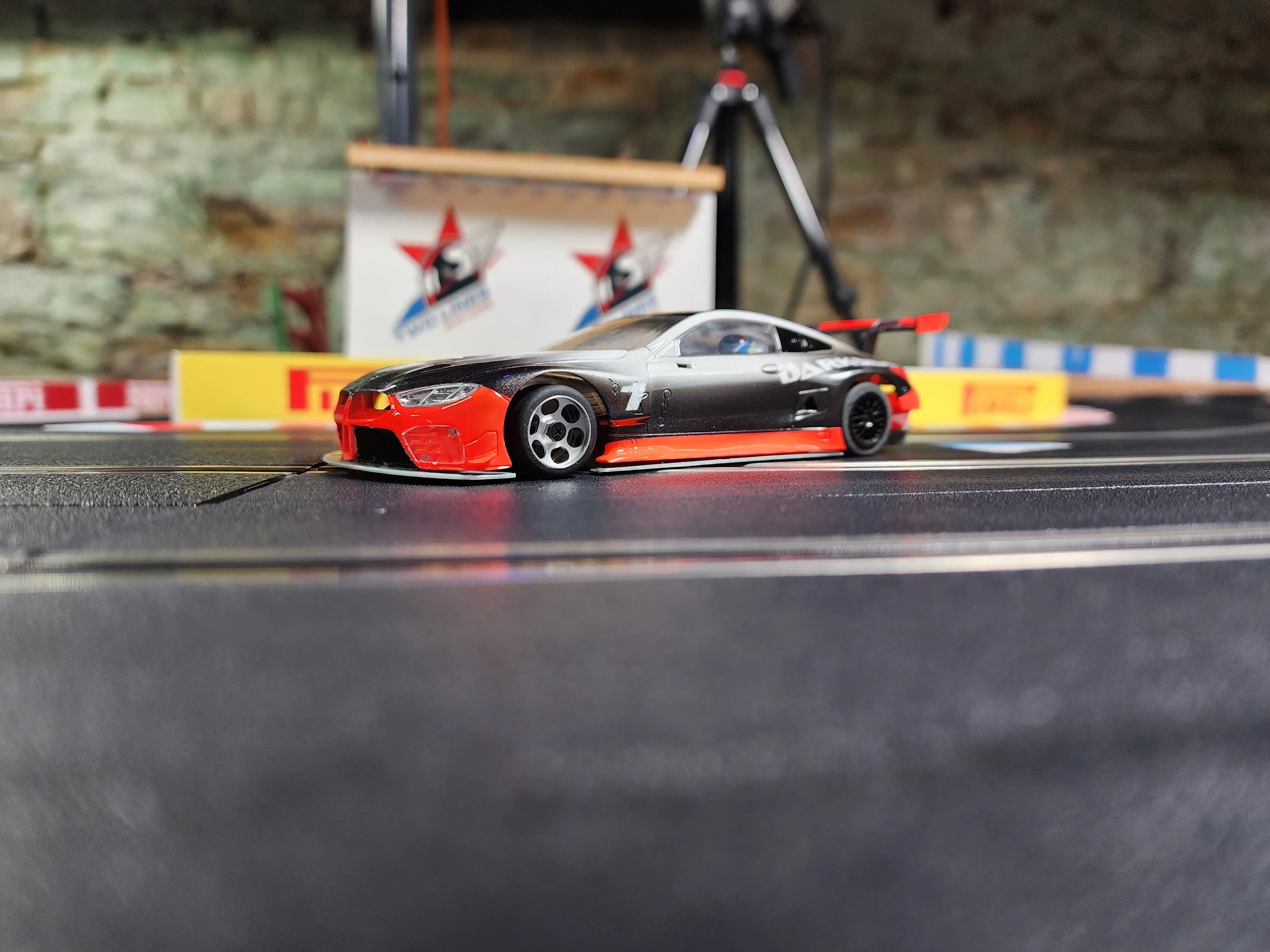 A detailed close-up of a black and red remote-controlled race car on a slot car racing track. In the background, there is a blurred poster with a logo, a camera tripod, and a stone wall.