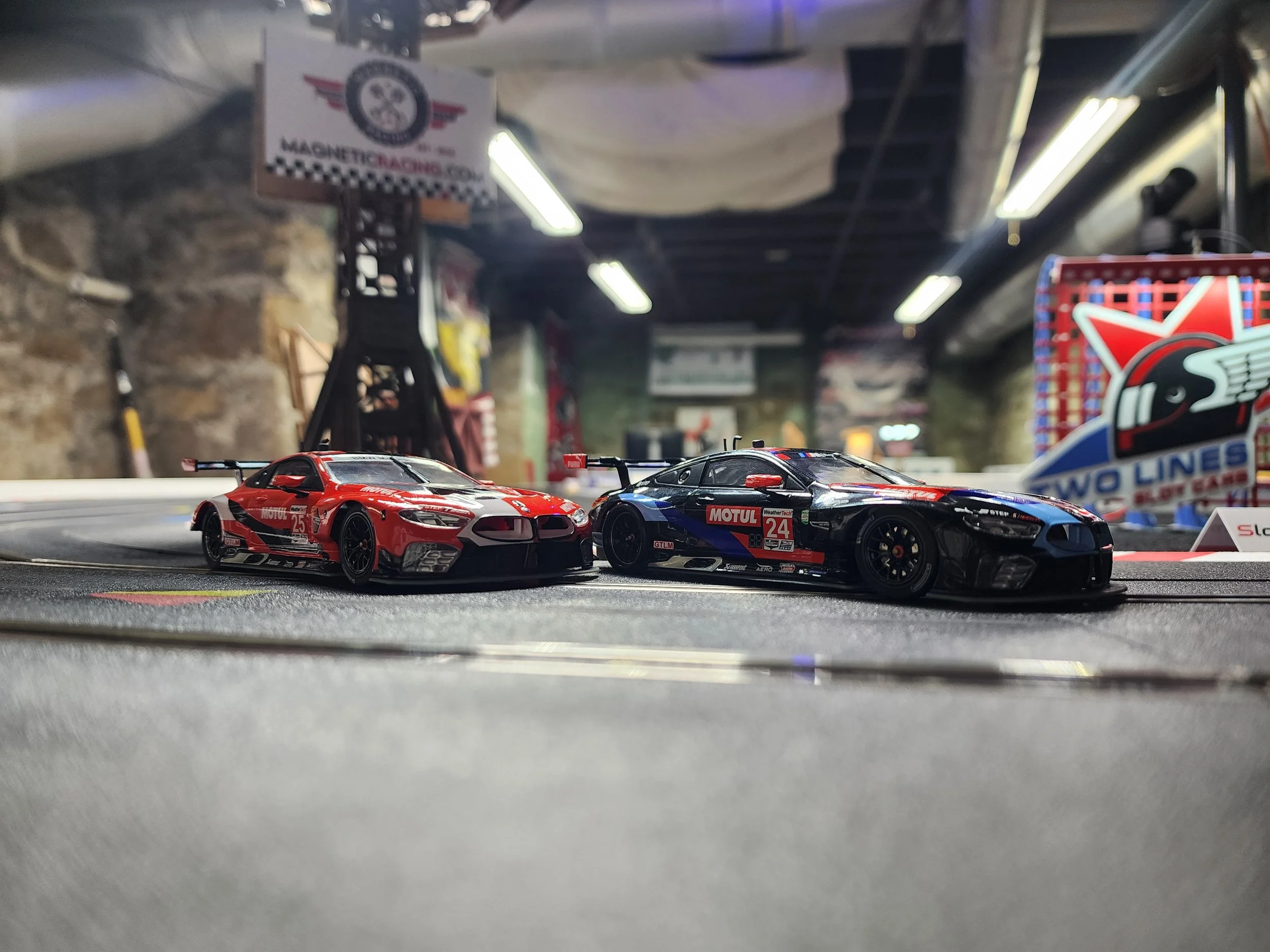 Two miniature race cars on a slot car racing track with a blurred background showing racing banners and a brick wall.