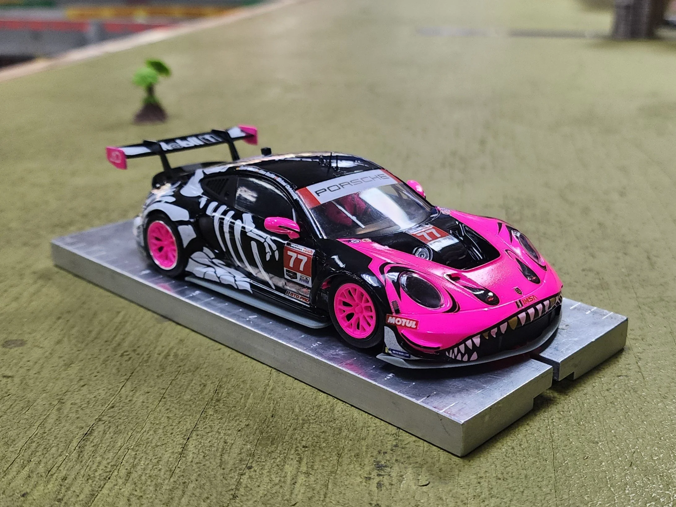 A detailed model of a pink and black Scaleauto Porsche 992 slot car on a metal platform, with a small green tree in the background on a green fabric surface.