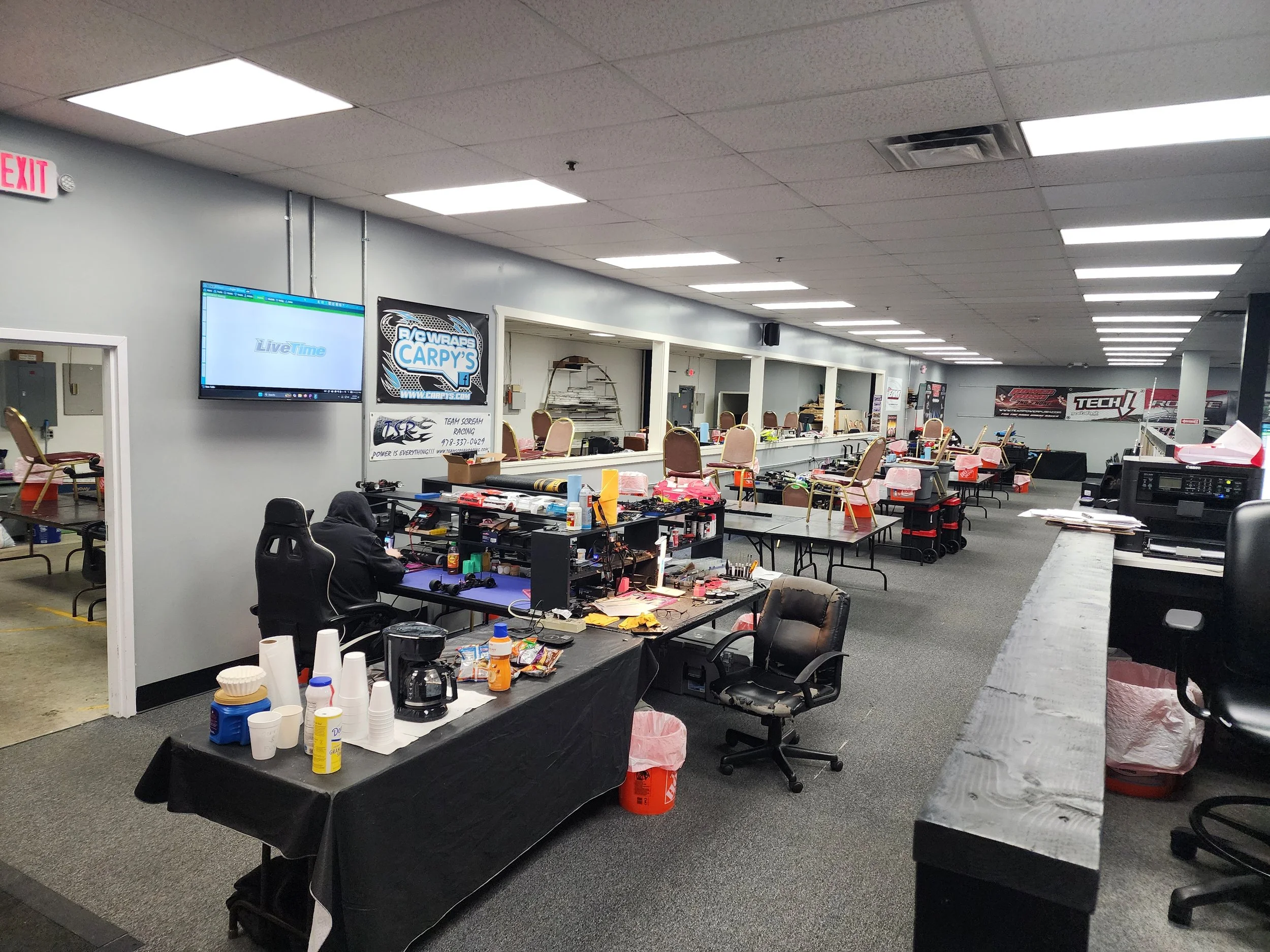 An indoor workspace with multiple tables, chairs, and electronic equipment, some tables have snacks and drinks. There is a TV and a banner on the wall, and a person is working at a desk.