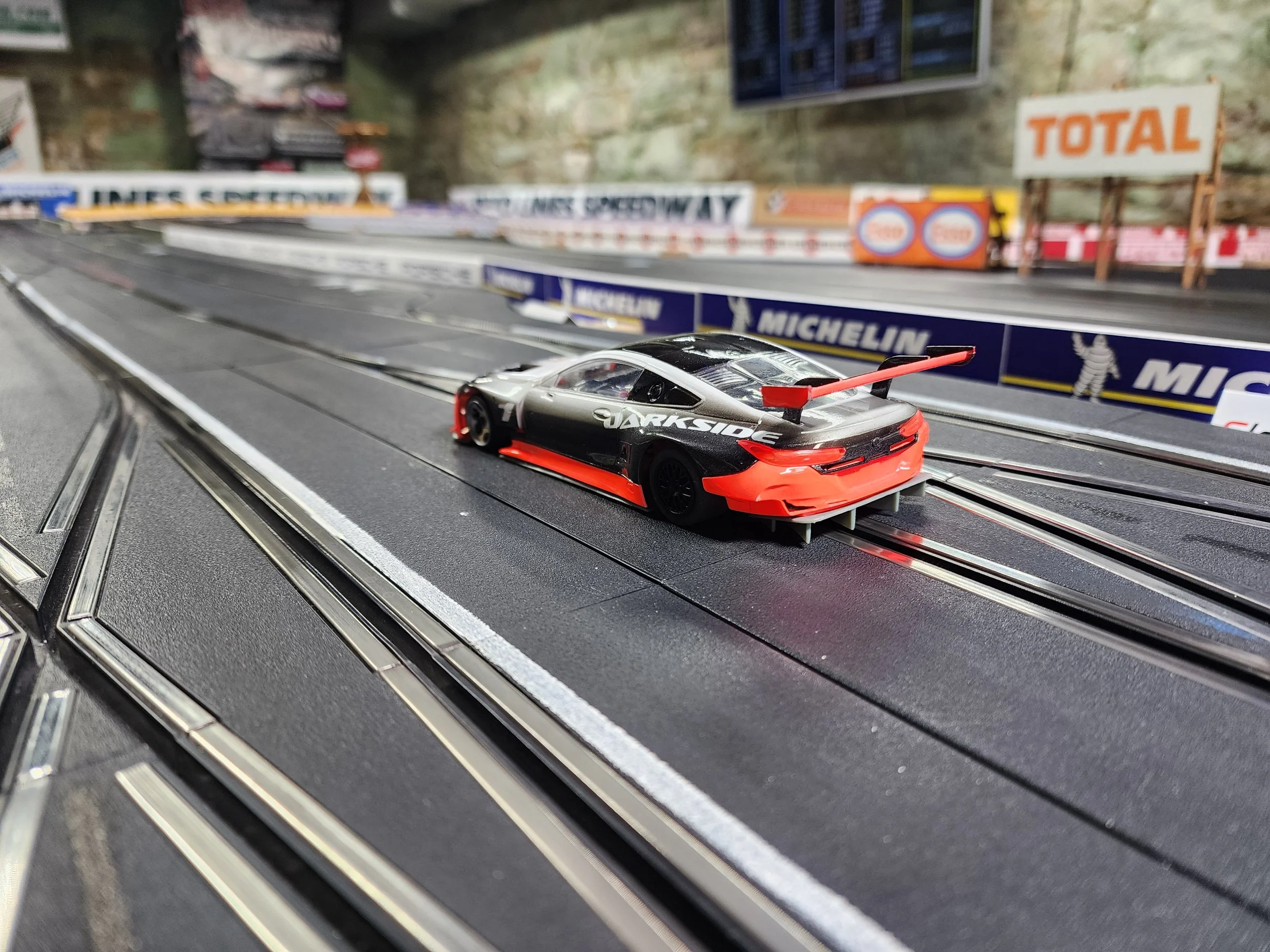 A miniature race car on a slot car racing track with branding and advertisements in the background.