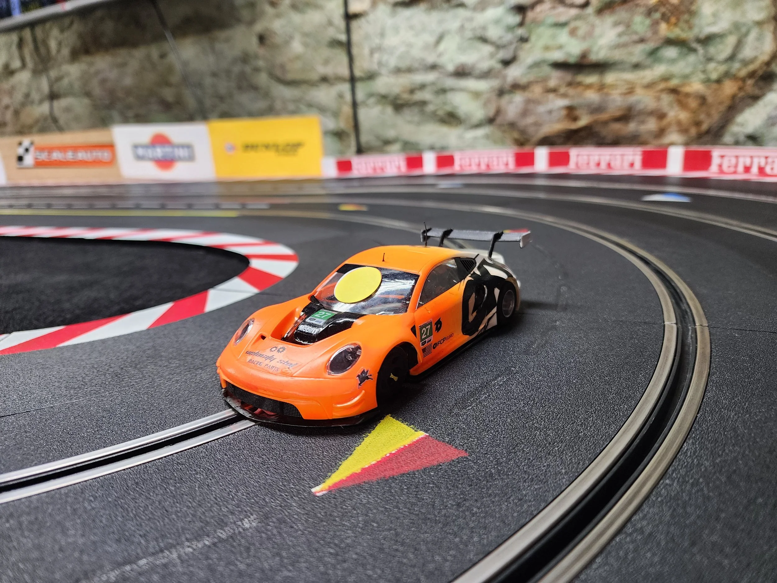 An orange slot car on a racing track with red and white curbs and a black and gray surface.