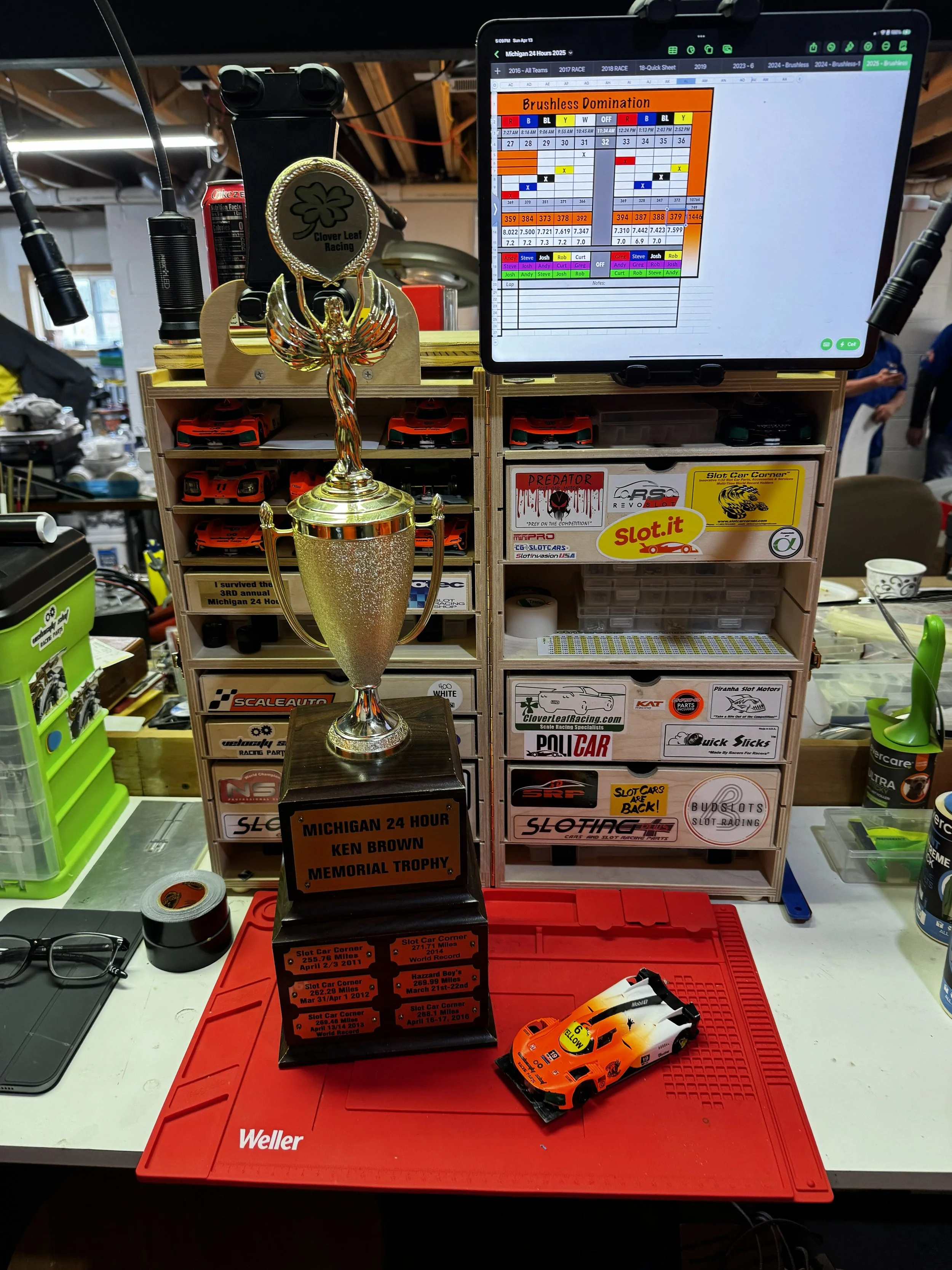 A model racing car trophy labeled 'Michigan 24 Hour Ken Brown Memorial Trophy' on a red desk mat with a small toy race car beside it. Behind the trophy, there are shelves with various toy cars, stickers, and a computer screen displaying a spreadsheet