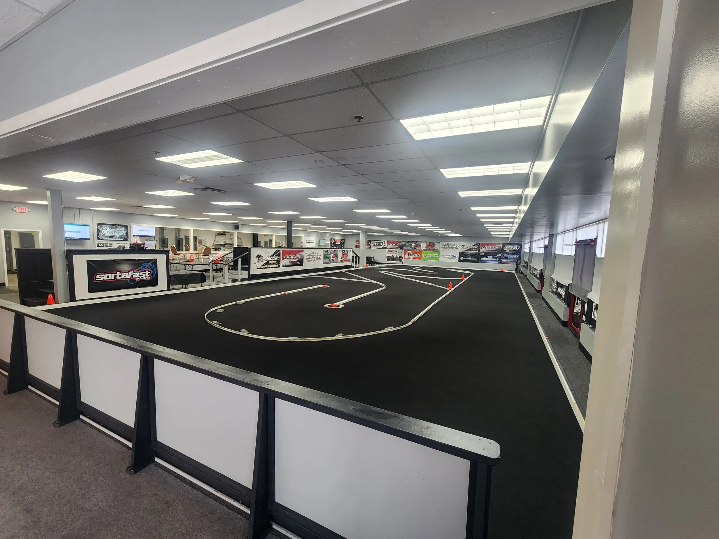 Indoor RC car racing track with black carpet surface, white barriers, and orange traffic cones, in a well-lit facility with banners on the walls.