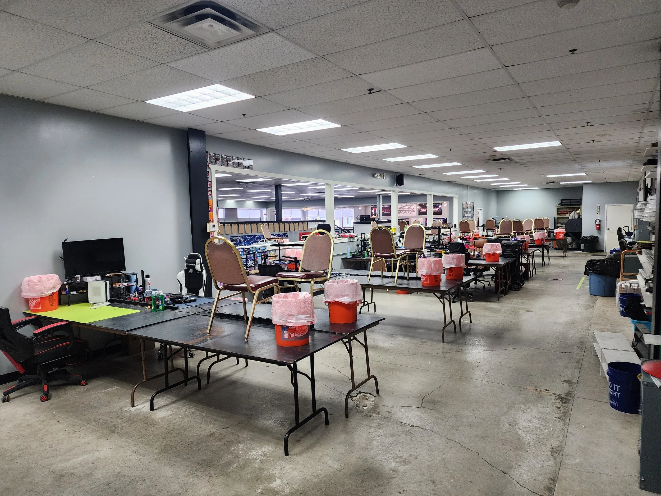A large indoor space with tables set up for an event, some with chairs on top, and various items such as buckets, chairs, and computers scattered around.