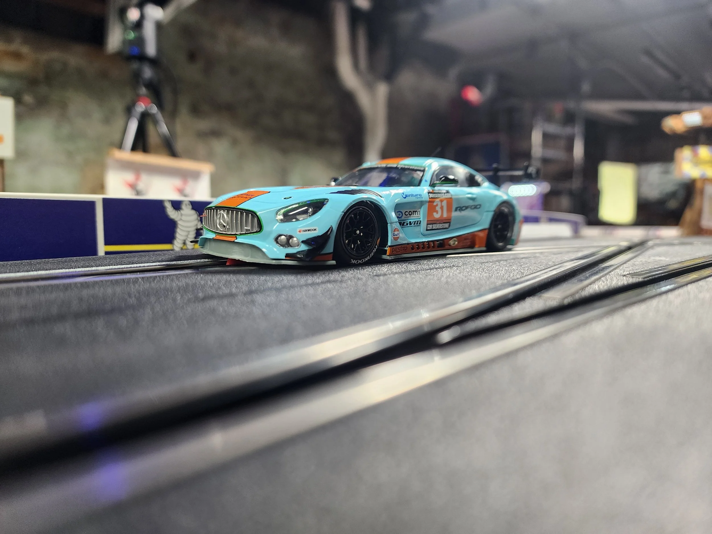 A miniature model race car on a slot car track with a blurred garage or hobby room background.