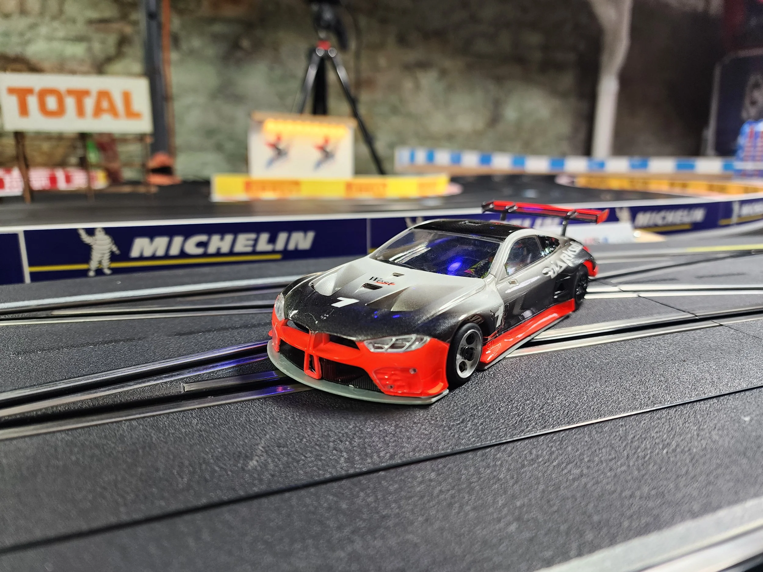 A detailed RC car model on a slot car racetrack with Michelin branding and racing posters in the background.