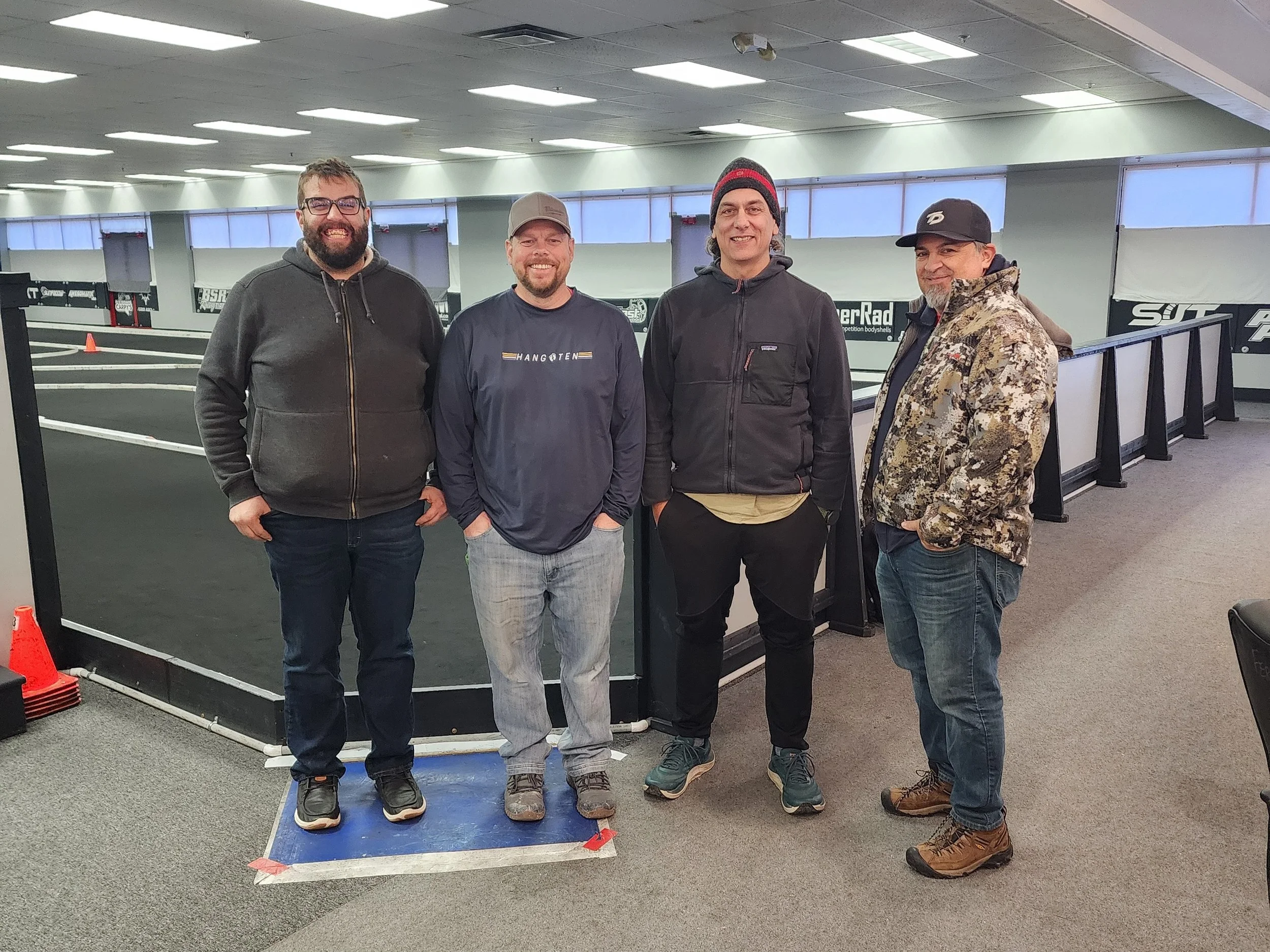 Four men standing inside an indoor R/C Car racing track, posing for a photo.