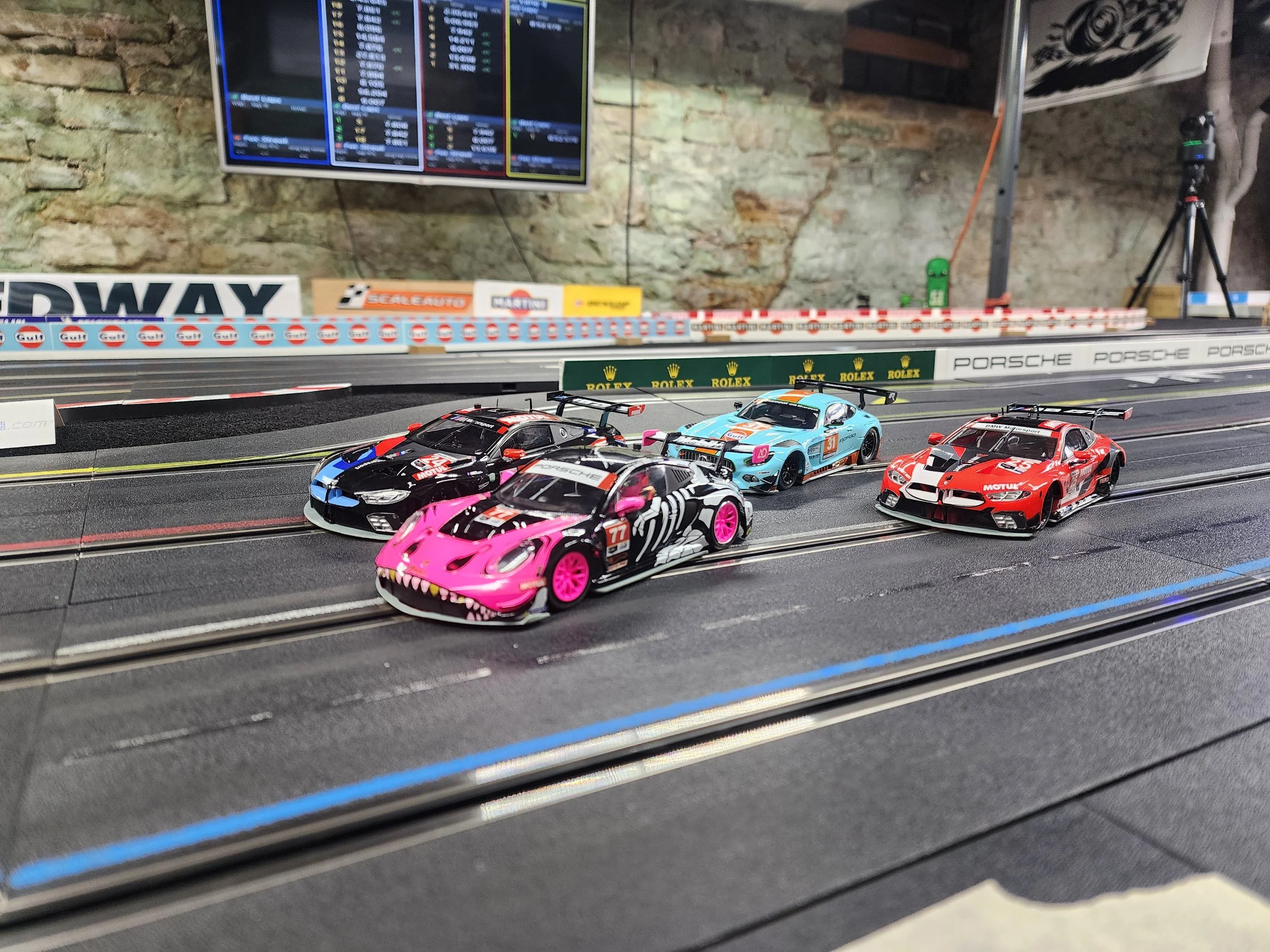 A slot car track with four slot cars, including BMW, Porsche, and other custom-designed cars, set against a backdrop of a stone wall, track advertisements, and a TV screen displaying race information.