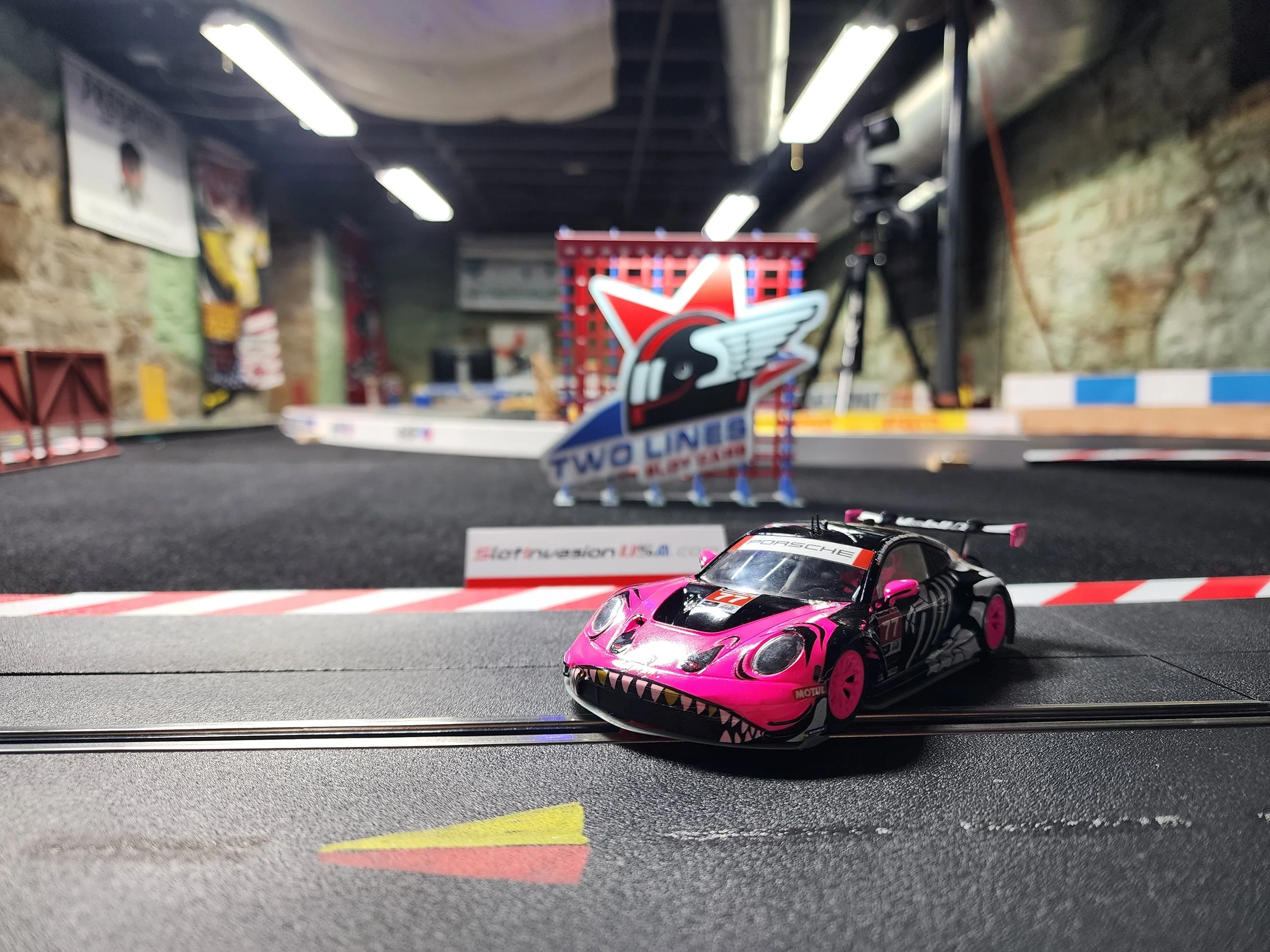 A pink and black slot car race car on a miniature race track with a large 'Two Lines' sign in the background.