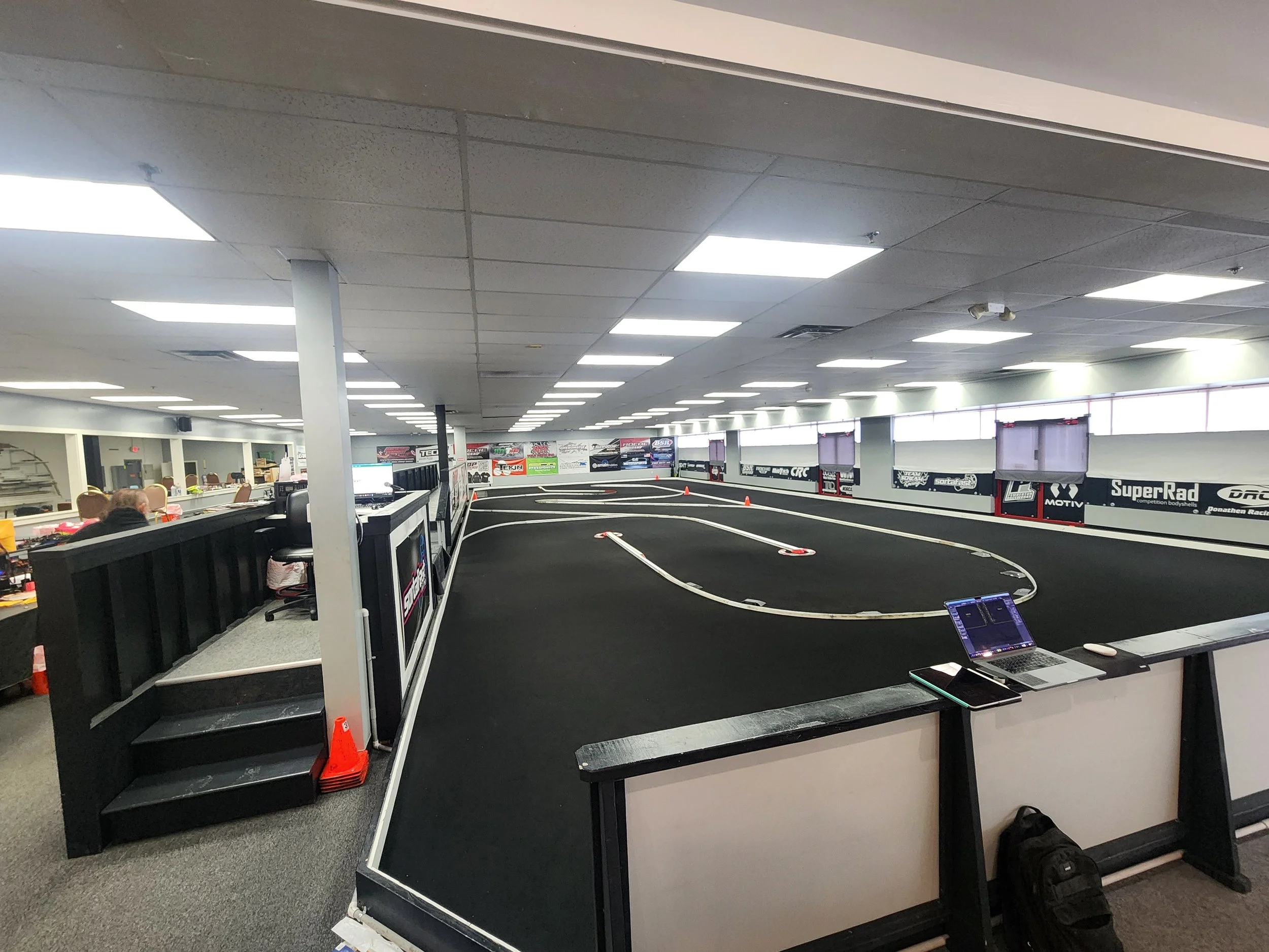 Indoor RC car racing track with black asphalt surface, white barriers, orange cones, and large electronic screens on the walls. There are tables and chairs on the left side, and a laptop and tablet on the foreground table.