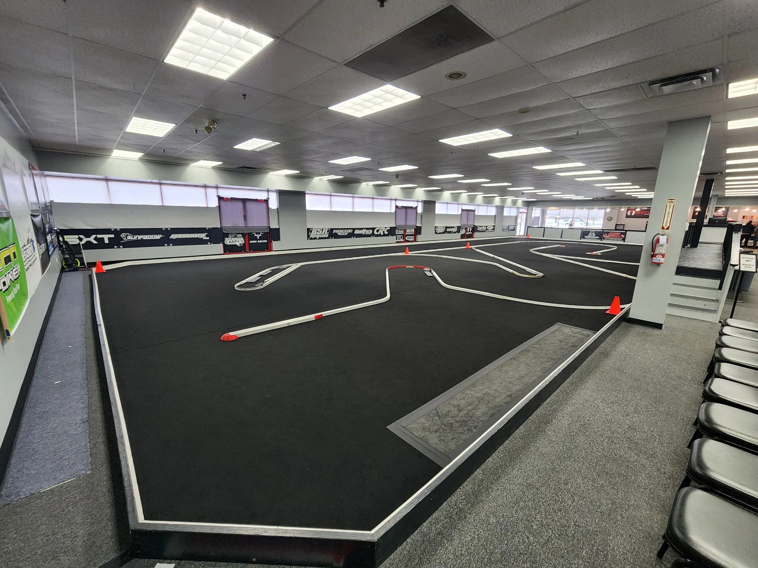 Indoor RC car racing track with black surface, white barriers, orange cones, and a staircase on the right side.