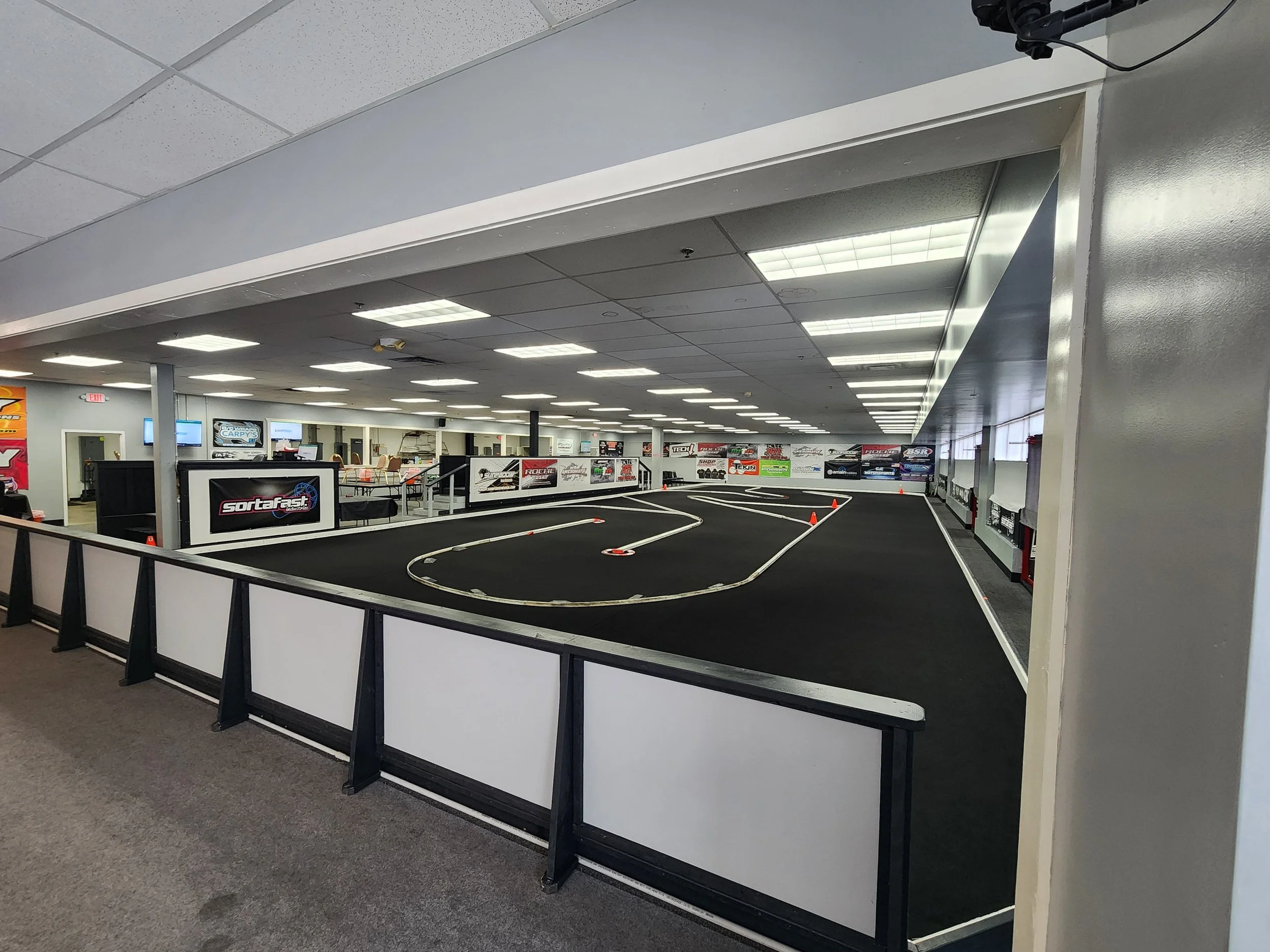 Indoor RC car race track with black surface, white track boundaries, orange cones, and surrounding seating and advertising banners.