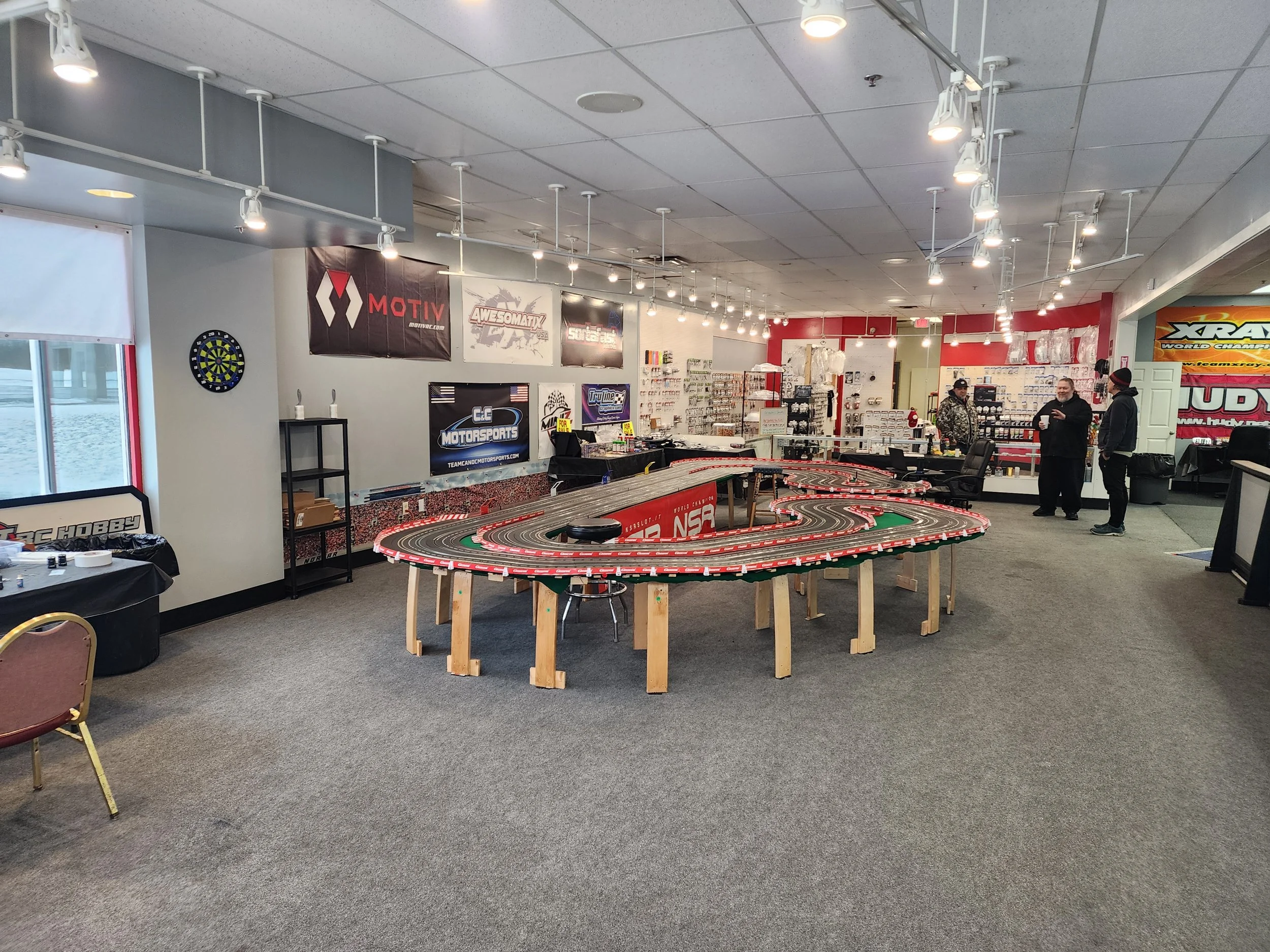 Interior of a hobby shop with a large slot car racing track in the center, surrounded by shelves displaying hobby supplies. Three people are standing and talking near the back, with banners and posters on the walls.