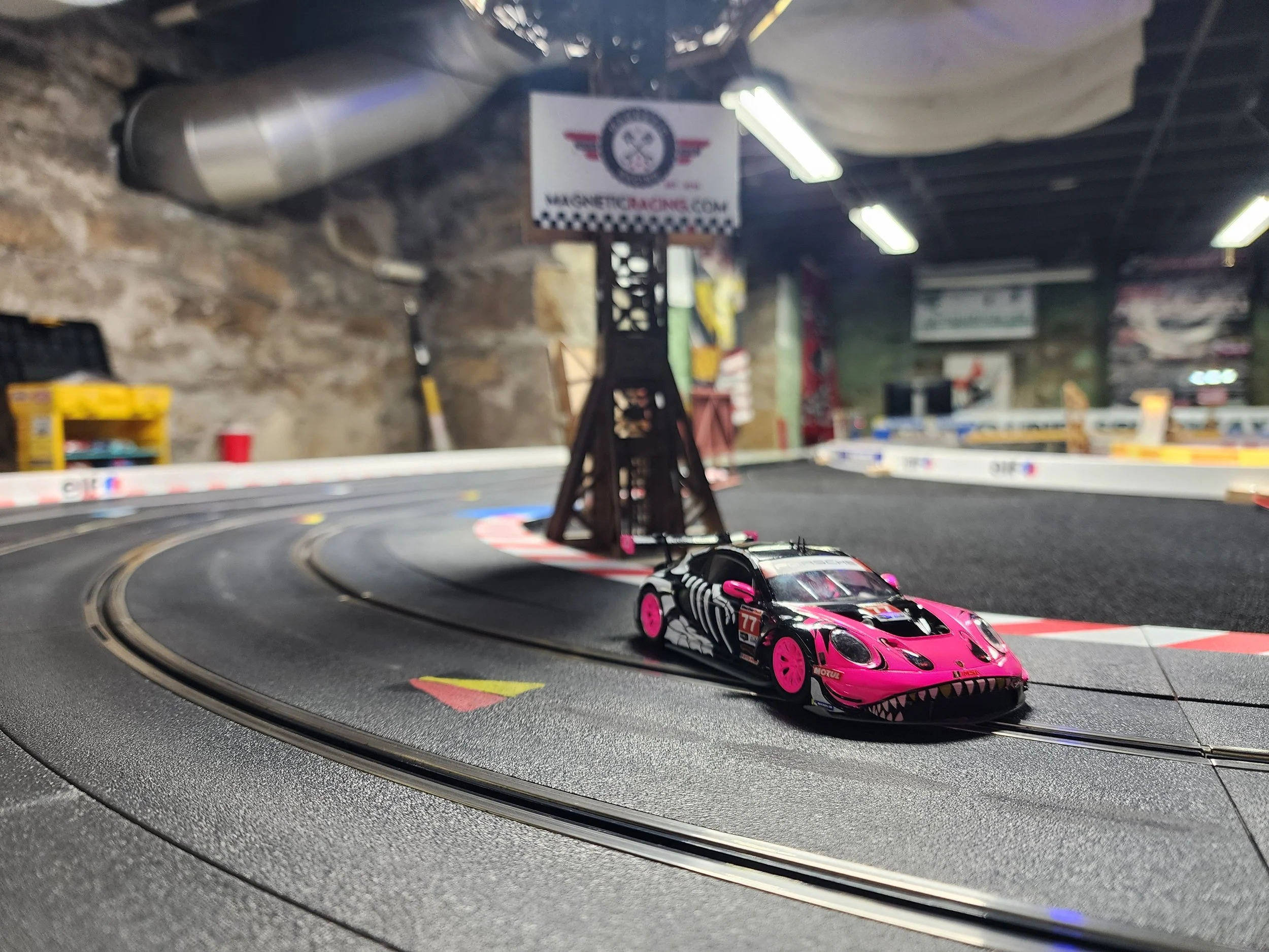 A pink and black slot car with tattoos and the number 77 on its side on a racing track in an arcade setting.