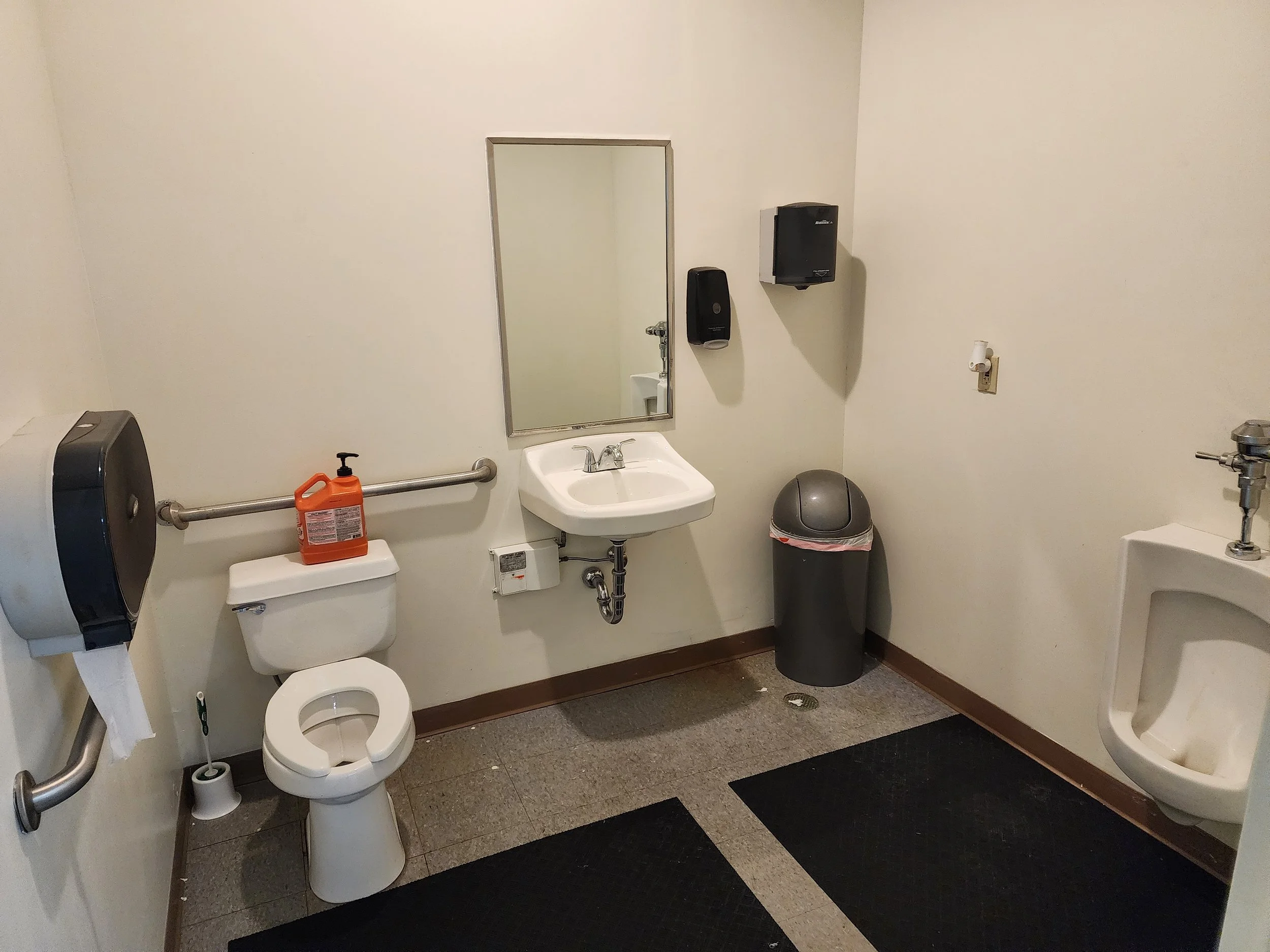 A public restroom with a toilet, a sink with a mirror above, a trash can, a urinal, and various paper towel dispensers.