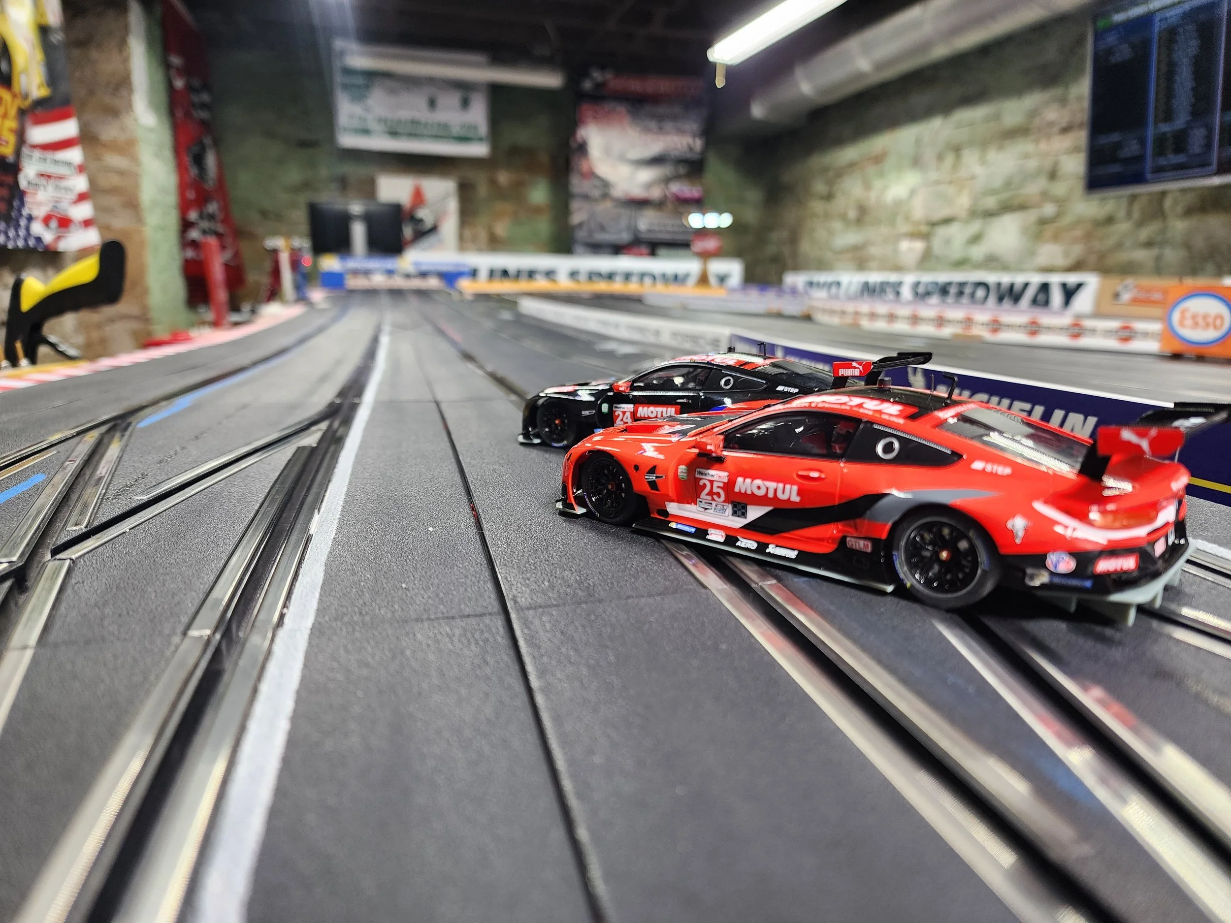 Close-up of two slot cars racing on a slot car track, with one red and black car in front and a black car behind, along a curved section of the track, with a blurry background of posters and banners.