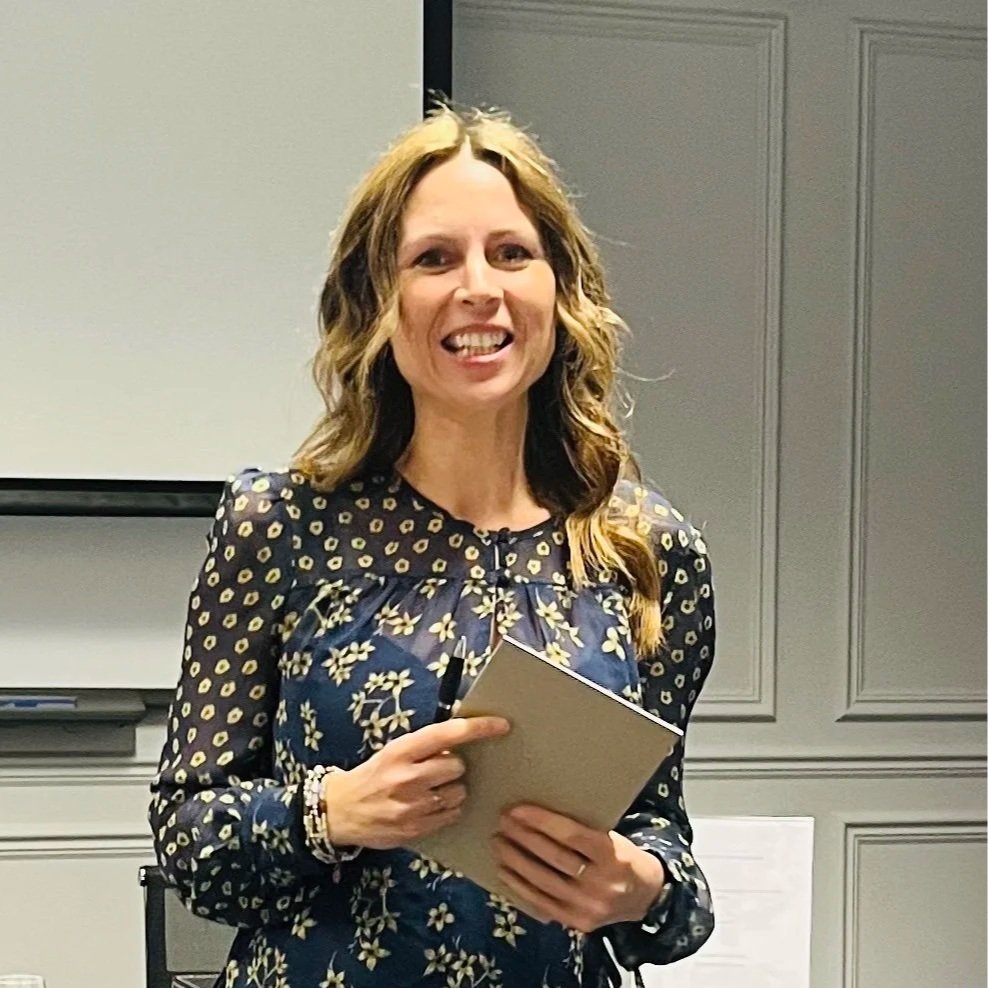 A woman with long, curly, blonde hair wearing a dark blue dress with a yellow floral pattern, holding a notebook, smiling in a room with gray paneled walls.