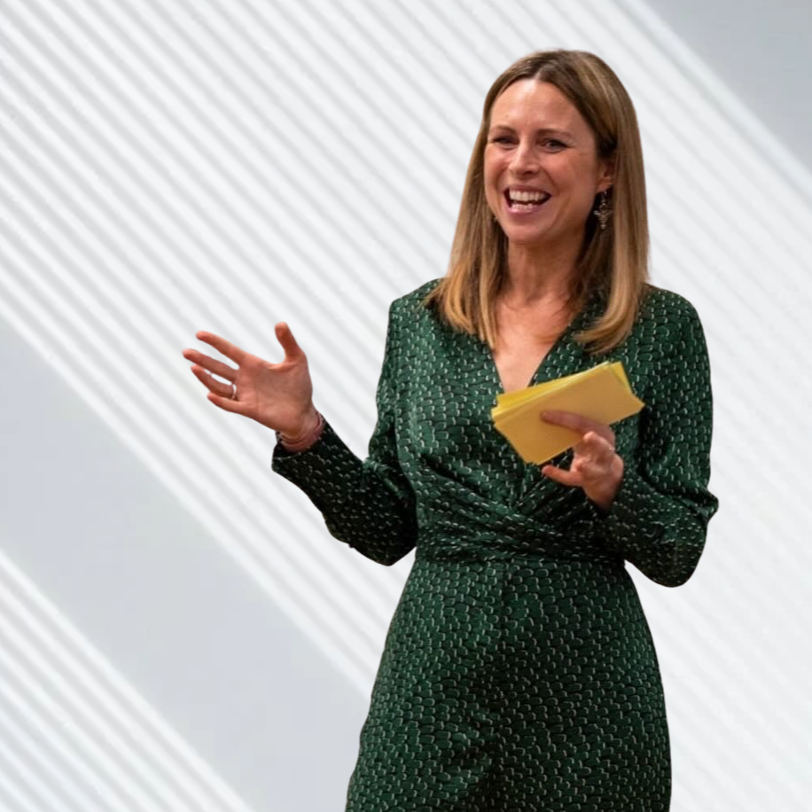 A woman with shoulder-length light brown hair, wearing a green patterned dress, holding yellow papers, smiling and speaking.