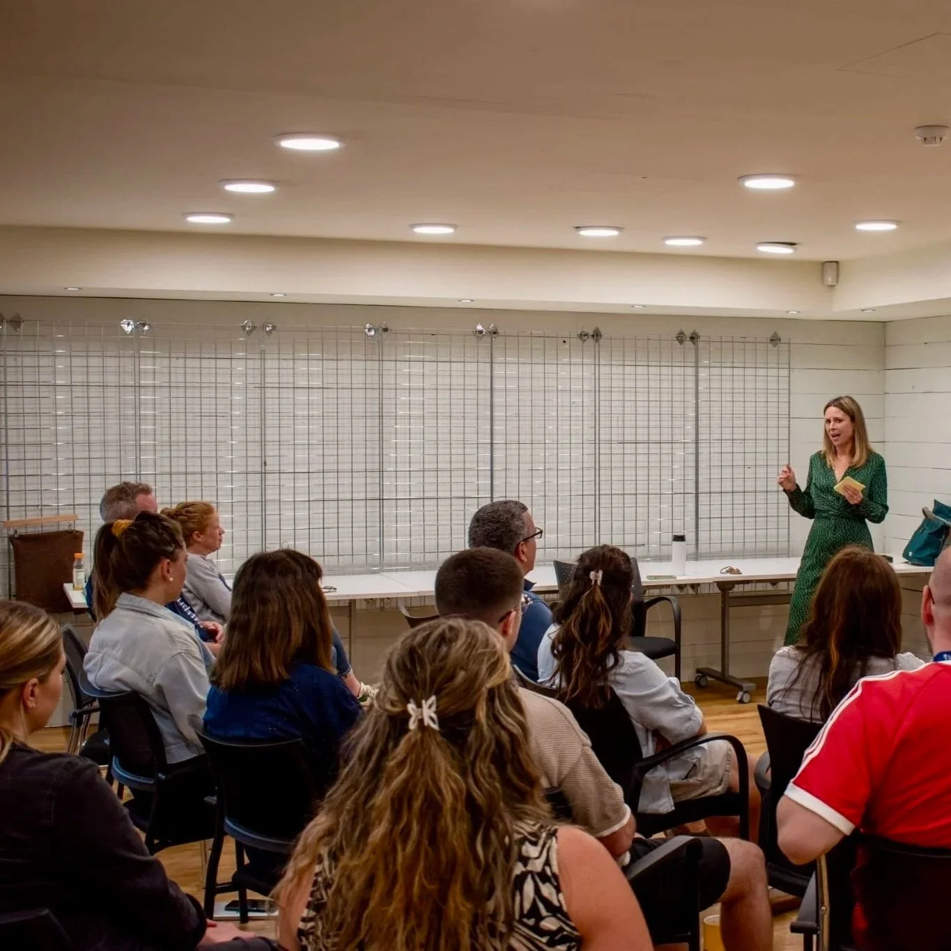 Woman giving a presentation to an audience in a room with white walls and a blank grid backdrop.