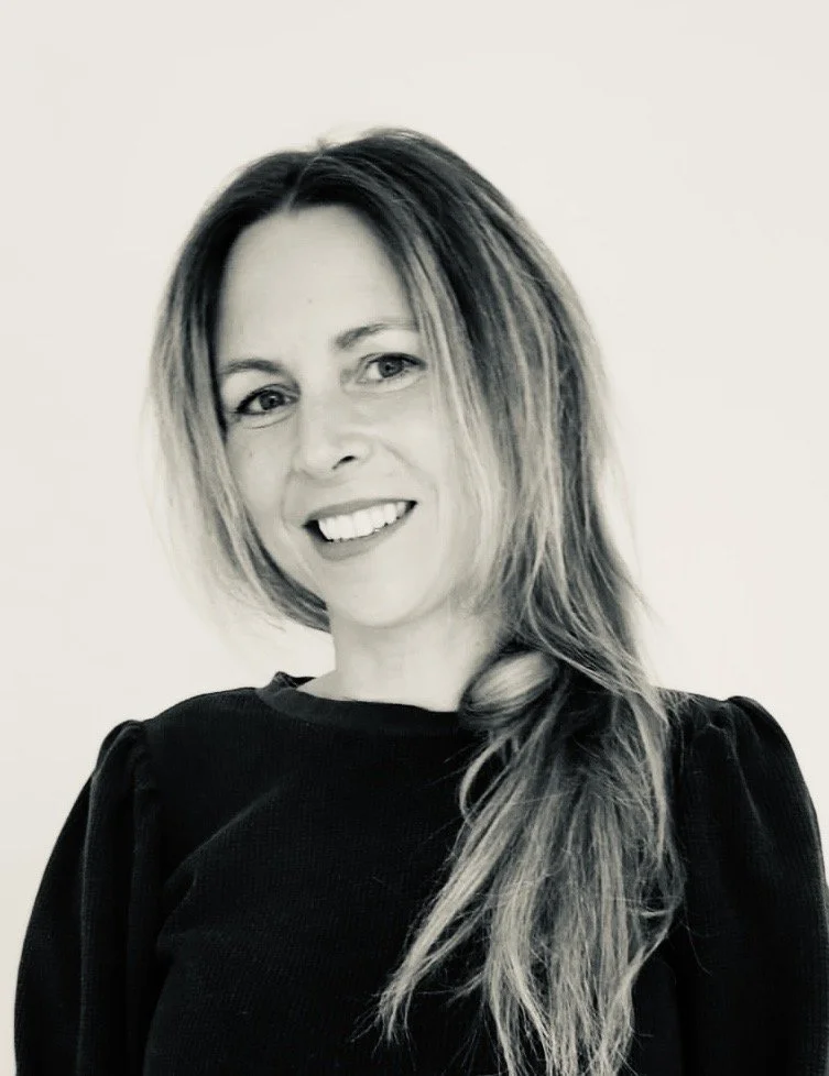 A woman with long, layered hair smiling at the camera, wearing a dark top, indoors with a plain light-colored background.