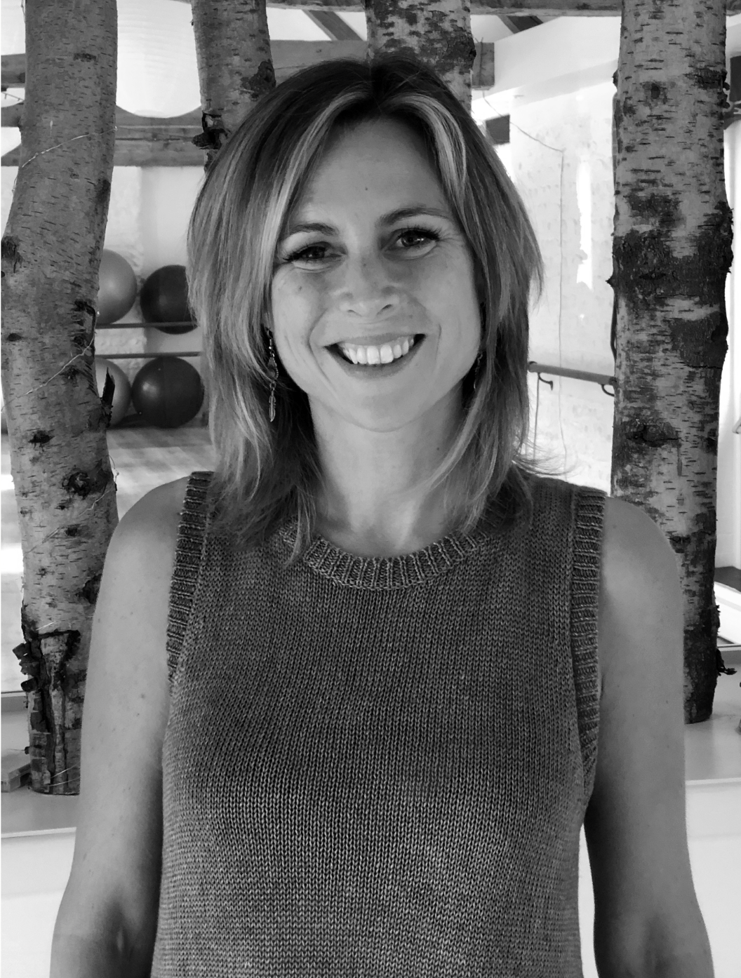 A woman with shoulder-length hair smiling, wearing a sleeveless knit top, standing indoors in front of trees and gym equipment.
