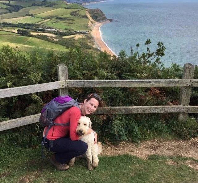 A woman with a purple backpack crouching next to a golden retriever puppy near a wooden fence with a coastal landscape in the background.