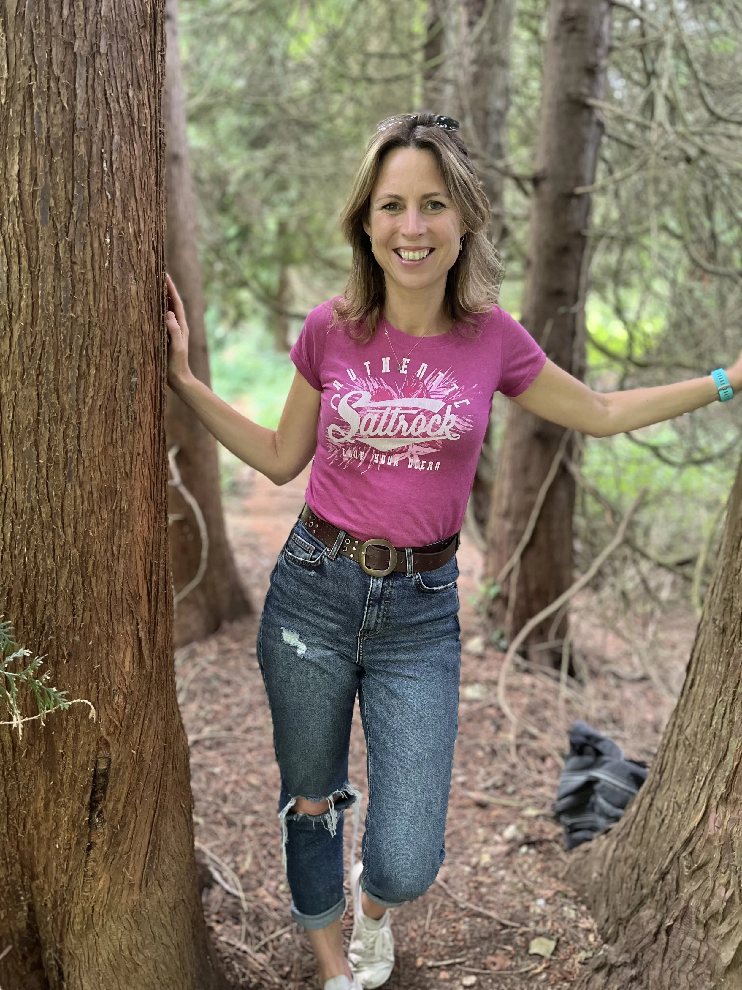 A smiling woman in a pink t-shirt and ripped jeans standing in a forest, holding onto trees on either side.
