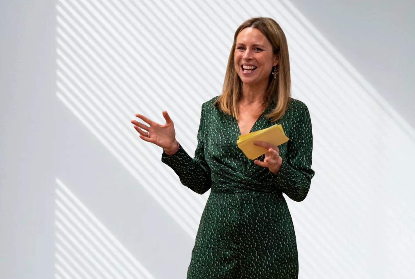A woman with shoulder-length brown hair smiling while speaking, holding a yellow notepad, wearing a green patterned dress, in front of a plain wall with light stripes.