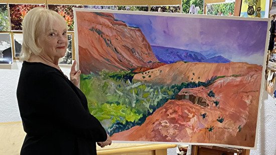 A photo of artist Sandra Humphries standing next to a large landscape painting of cliffs, mountains, and greenery.