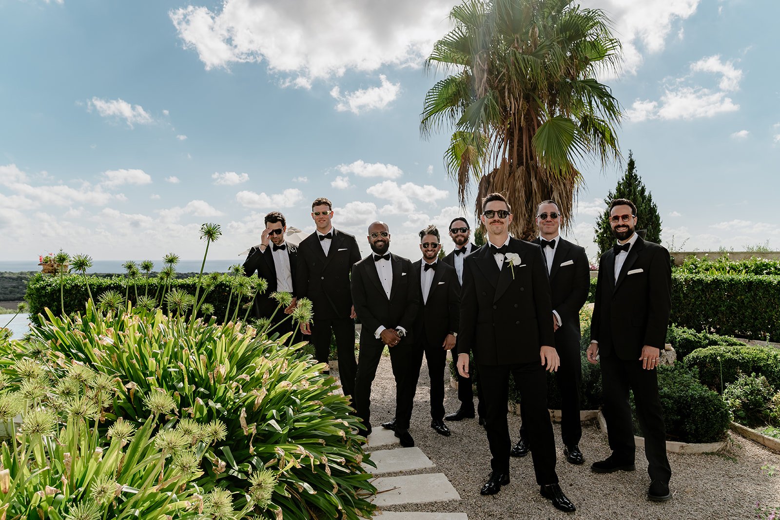 Group of groomsmen in black tuxedos posing in a Mediterranean garden overlooking the sea in Menorca